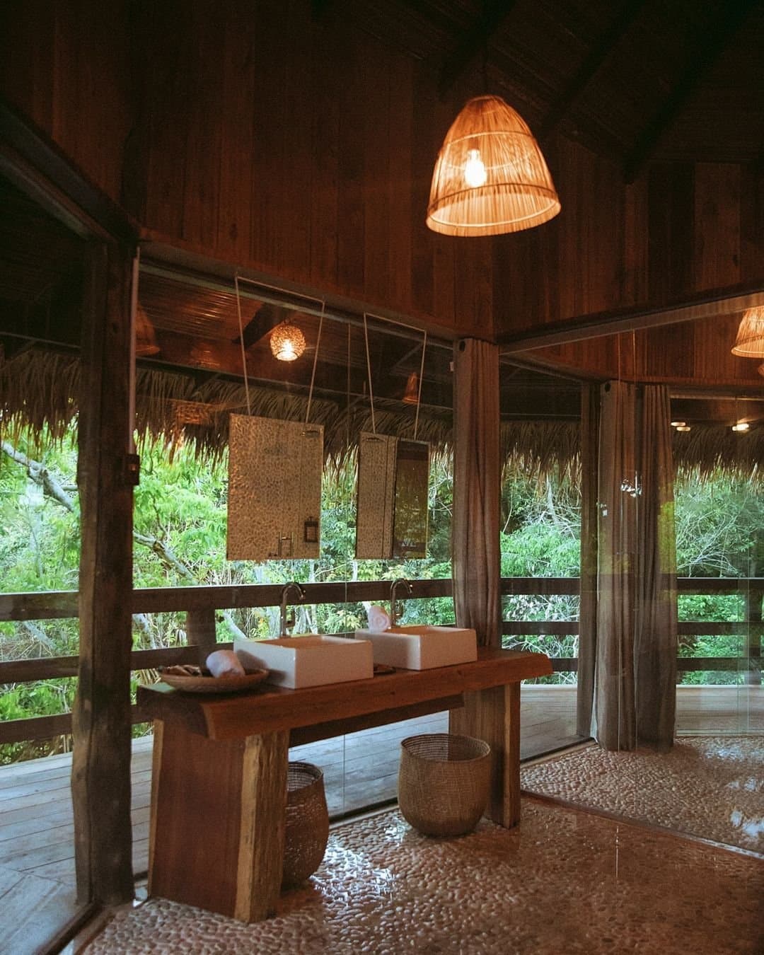 Duschen mit Regenwaldsicht, Luxus, der sich ganz natürlich anfühlt. Offenes Badezimmer in der Juma Amazon Jungle Lodge mit zwei Aufsatzwaschbecken auf einem Massivholz-Waschtisch, geflochtenem Hängelampenschirm, Kieselsteinboden und raumhohen Glaswänden mit Blick in den Regenwald