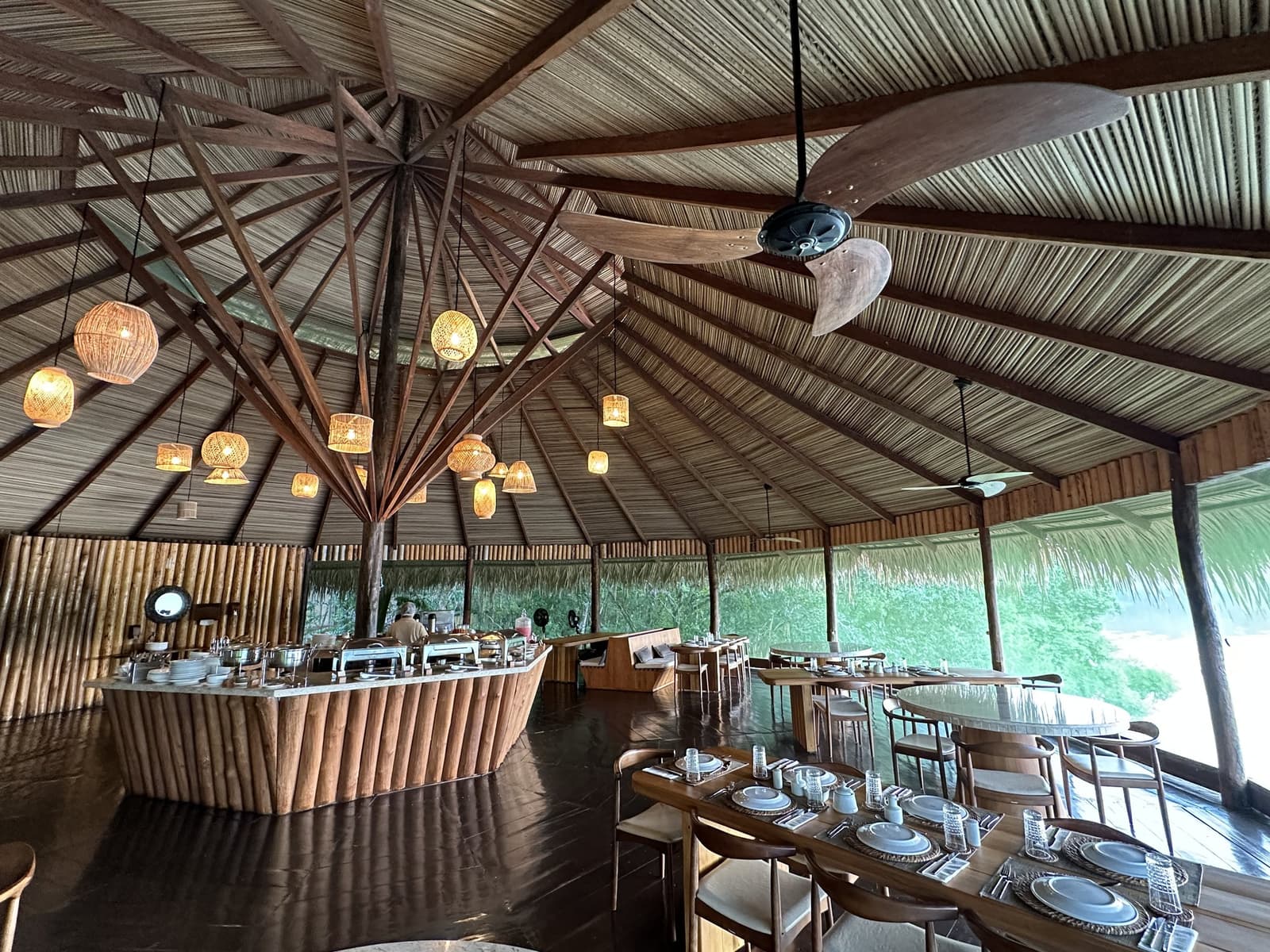 Essen unter Palmblättern, während der Regenwald neugierig hereinschaut. Das Open-Air-Restaurant des Juma Amazon Jungle Lodge mit einem hohen Strohdach aus radialen Holzbalken, geflochtenen Rattanleuchten, einer bambusverkleideten Buffettheke und gedeckten Holztischen, umgeben von üppigem Regenwald