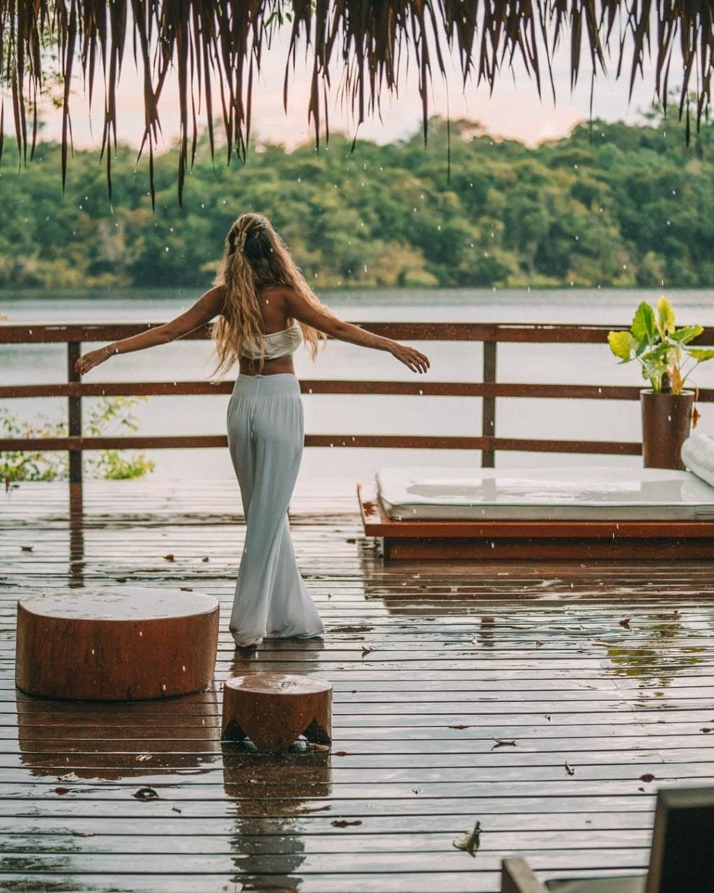 Warmer Regen, weite Arme, kein Plan. Genau so. Eine Frau in Weiß steht mit ausgebreiteten Armen auf einer regenfeuchten Terrasse der Juma Amazon Jungle Lodge und blickt auf einen ruhigen Fluss mit dichtem Regenwaldufer