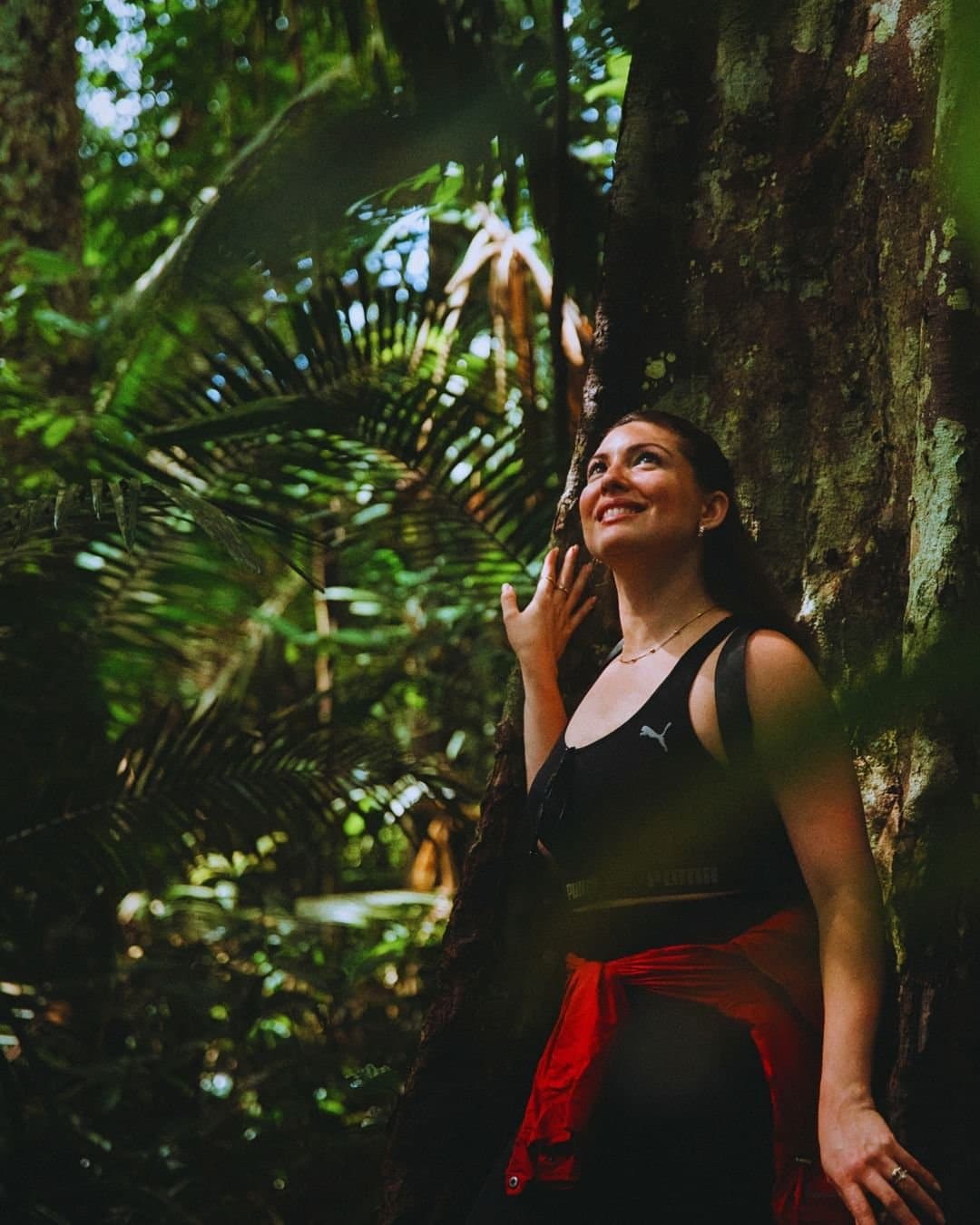 Manche Bäume lassen einen einfach innehalten und staunen. Eine lächelnde Frau lehnt auf einer geführten Wanderung des Juma Amazon Jungle Lodge an einem riesigen Regenwald-Baumstamm, umgeben von üppigen Farnen und hohen Palmen