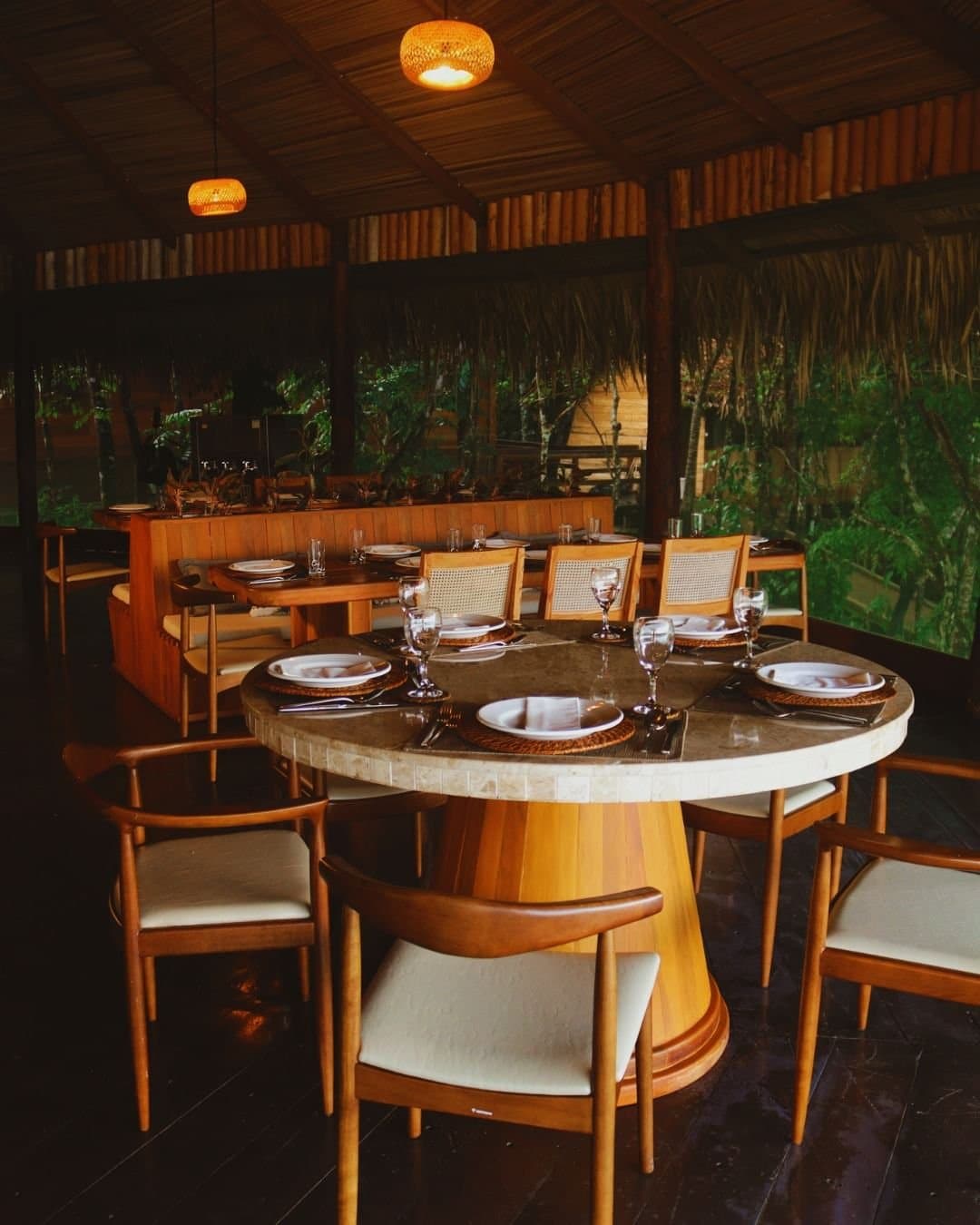 Abendessen im Regenwald, wo der Urwald fast schon am Tisch sitzt. Gedeckter ovaler Esstisch mit geflochtenen Untersetzern, Weingläsern und weißen Tellern im offenen Restaurant der Juma Amazon Jungle Lodge, umgeben von Bambuswänden, einem Strohdach und üppigem Regenwald