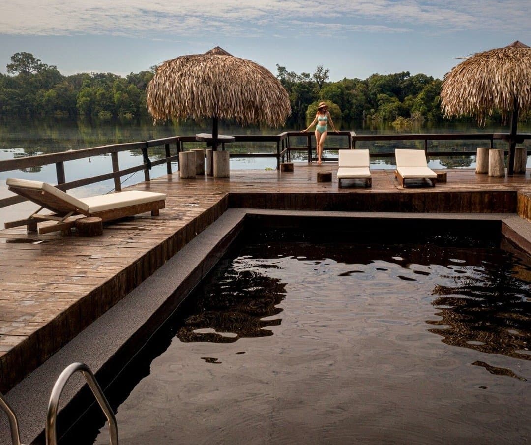 Schwarzwasser, Flussblick, keine Termine, das Amazonas-Tempo ist ansteckend. Ein Fluss-Pool mit dunklem Wasser auf einem Holzdeck der Juma Amazon Jungle Lodge, mit Strohdach-Sonnenschirmen, Liegestühlen und einer Gästin mit Blick auf den ruhigen Dschungelfluss
