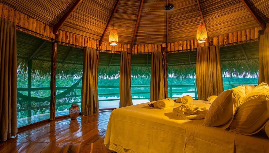 Schlafen mit Flussblick, aufwachen mit Vogelstimmen, so sieht ein guter Plan aus. Inneres eines Bungalows im Juma Amazon Jungle Lodge mit Doppelbett, geflochtenen Hängelampen, Reetdach und offenen Fenstern mit Blick auf einen ruhigen Dschungelfluss