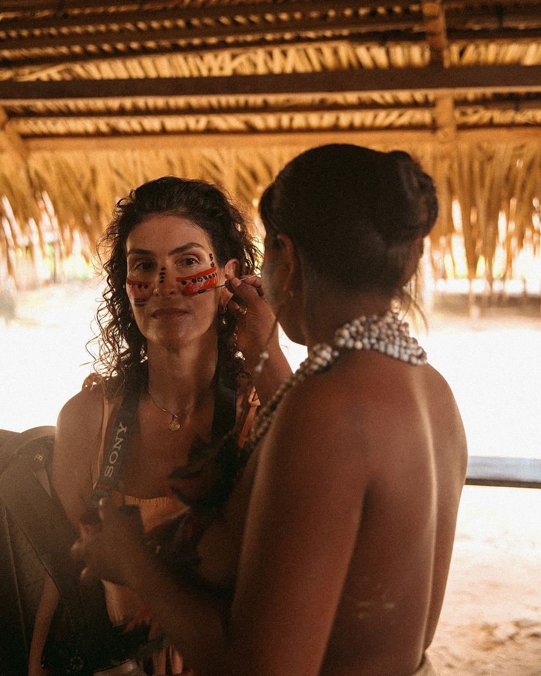 Ein stilles Gespräch, das keine Worte braucht. Eine indigene Frau trägt traditionelle rote und schwarze Gesichtsbemalung auf eine lächelnde Reisende in einer strohgedeckten Hütte im Juma Amazon Jungle Lodge auf