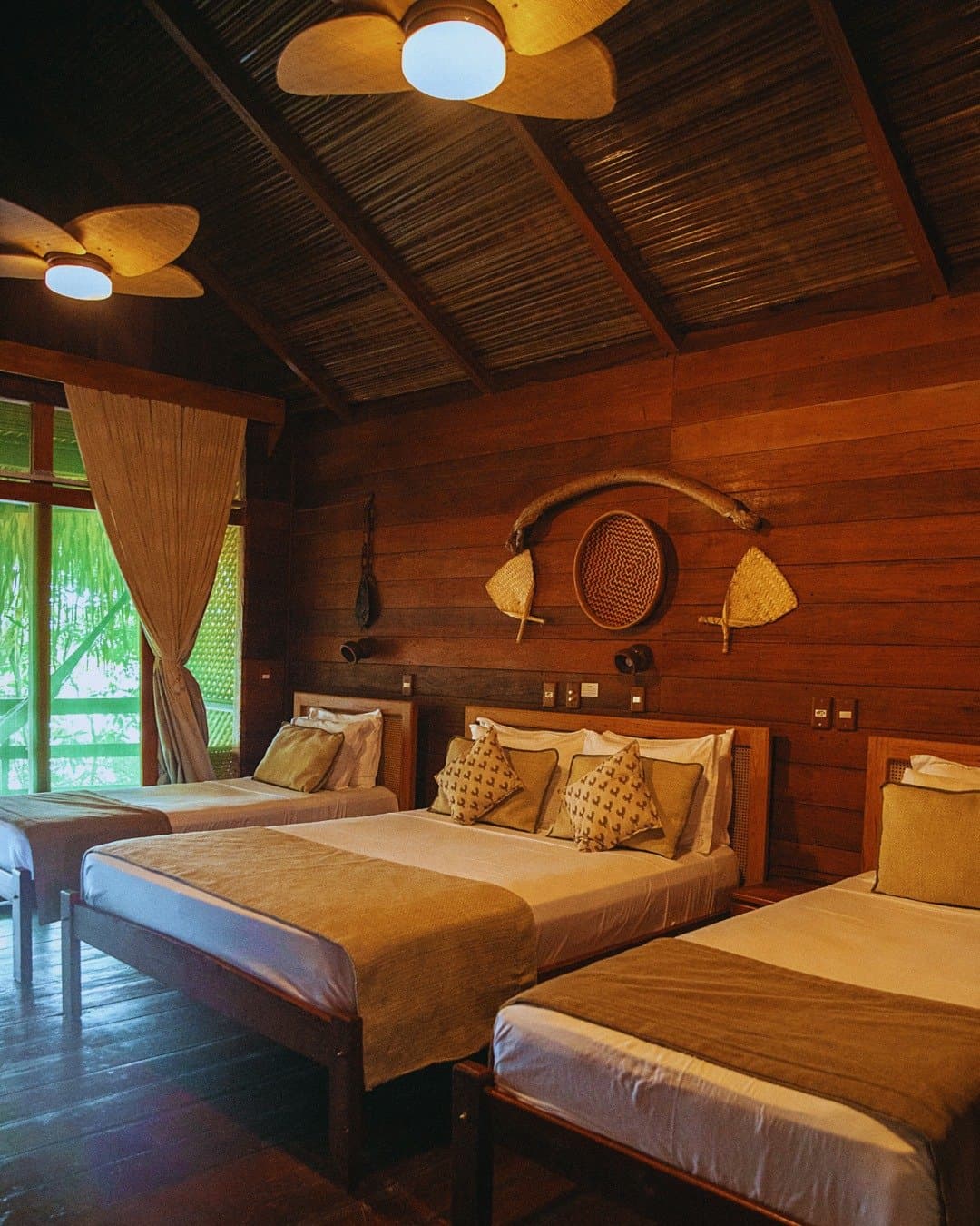 Geflecht, Holz und Regenwald vor dem Fenster, Schlafkomfort mit echtem Charakter. Dreibettzimmer in der Juma Amazon Jungle Lodge mit drei Holzbetten, gewebten Wanddekorationen, einem Strohdach mit Deckenventilatoren und Blick auf den Regenwald durch die Balkontür
