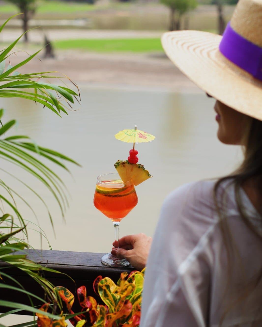 Sundowner mit Seeblick, und der Dschungel gehört dazu. Eine Frau mit einem breitkrempigen Strohhut hält im Juma Amazon Jungle Lodge einen leuchtend orangen Cocktail mit Ananas, Kirsche und Papierschirmchen, dahinter ein ruhiges Gewässer und üppiges Grün