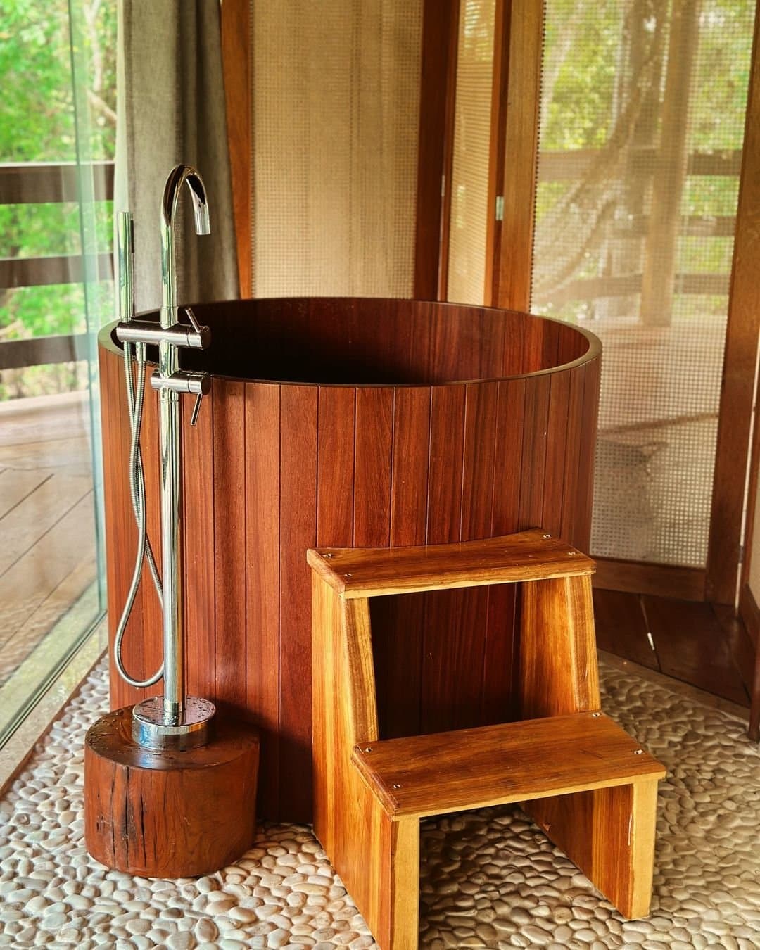Baden im Urwald, der Regenwald schaut einfach durchs Fenster zu. Rundes poliertes Holz-Badebecken mit verchromtem Standarmatur und Holztritthocker auf Kieselsteinboden, mit Waldsicht-Fenstern im Juma Amazon Jungle Lodge