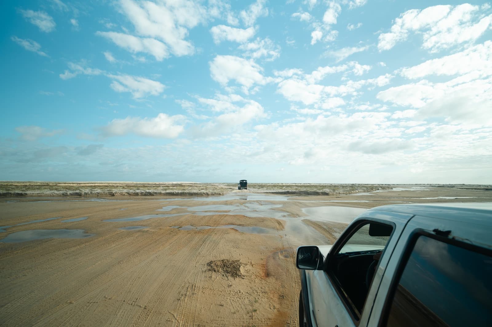 Het getij trekt zich terug en laat een route achter. Twee terreinwagens rijden over een uitgestrekte natte zandvlakte onder een brede blauwe lucht tijdens een Lençóis Maranhenses trip, met ondiepe plassen die het licht weerkaatsen