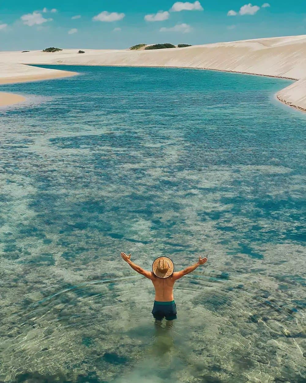 Armen wijd, hoofd leeg, precies wat je nodig hebt. Een reiziger staat tot aan de heupen in een kristalheldere turquoise lagune tijdens een Lençóis Maranhenses trip, armen wijd gespreid, met witte zandduinen die zich uitstrekken onder een stralend blauwe hemel