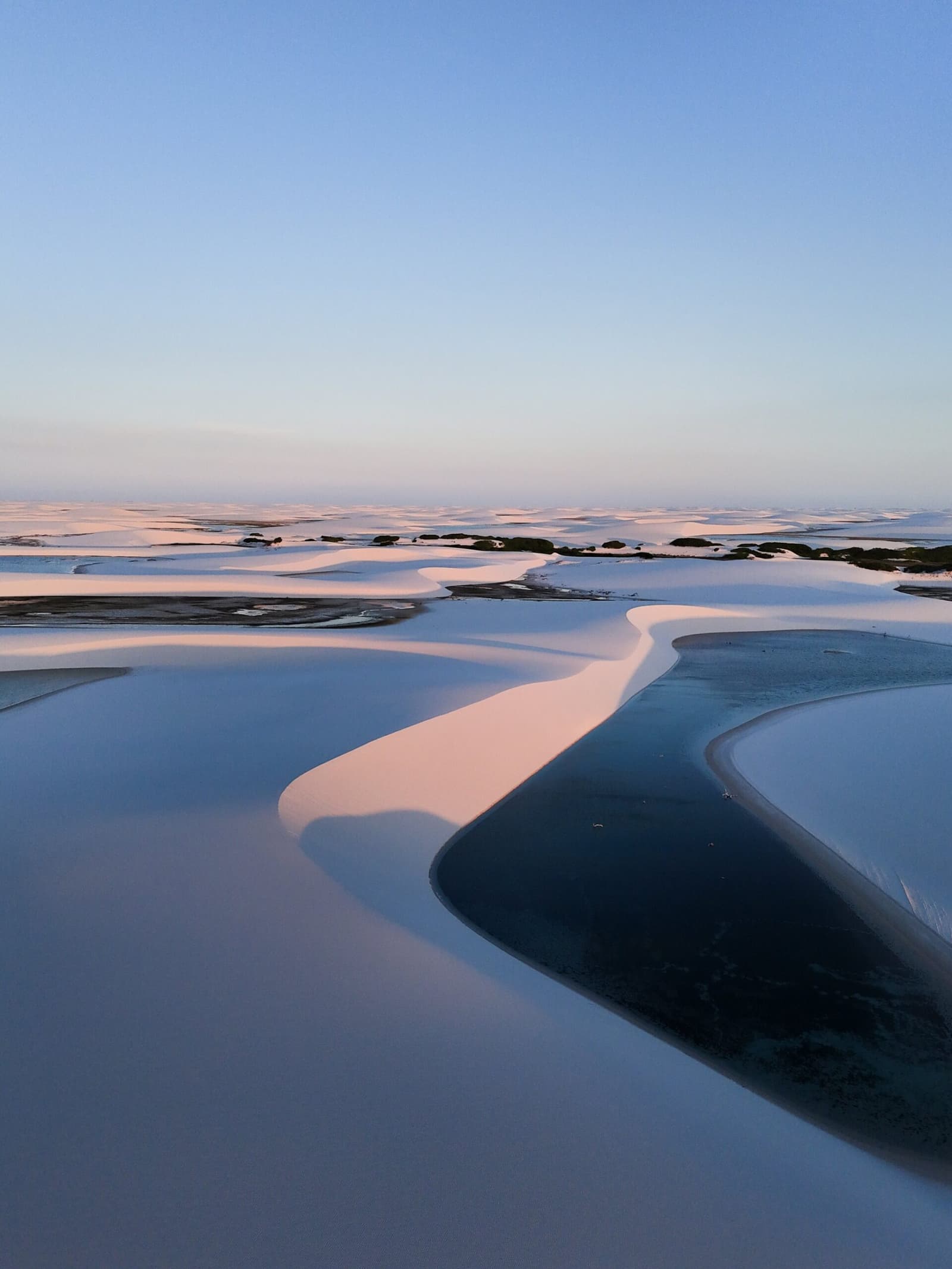 Roze licht, wit zand, koud water, de Lençóis op hun stilste en mooiste. Luchtfoto van witte zandduinen en donkerblauwe lagunes die roze gloeien onder een schemerende hemel tijdens een Lençóis Maranhenses-reis