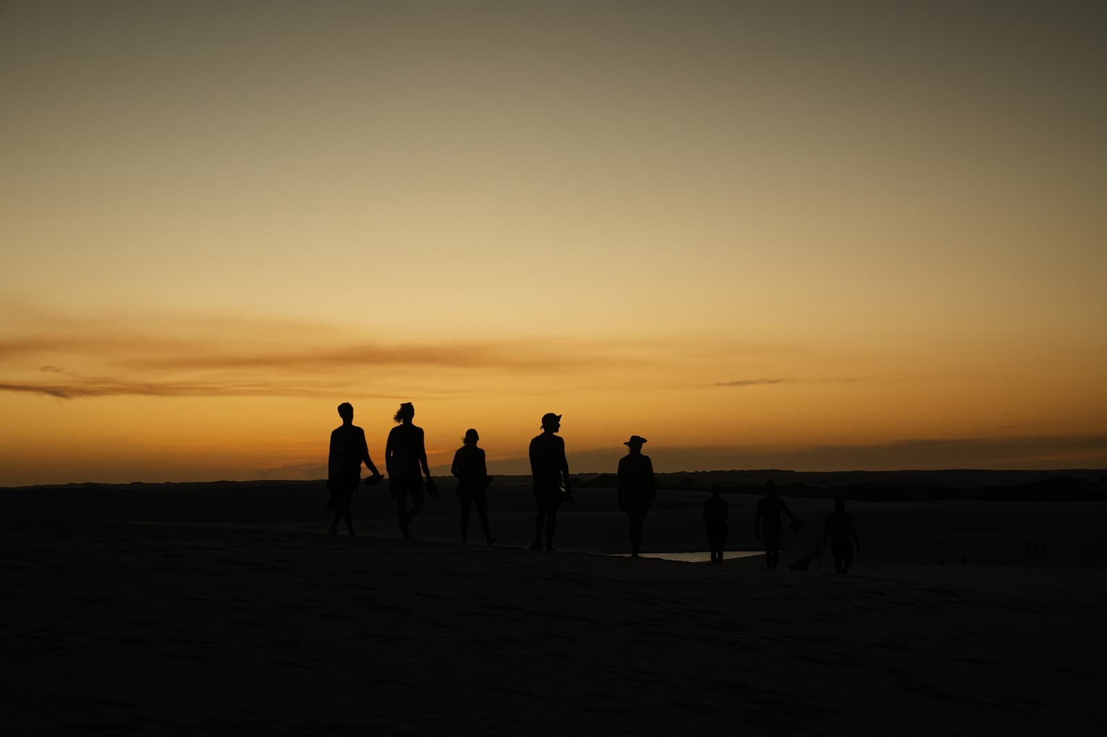Op dit moment weet iedereen precies waarom ze zijn gekomen. Silhouetten van een groep reizigers die over open duinen lopen tijdens een Lençóis Maranhenses-trip, afgetekend tegen een vurig oranje avondlucht