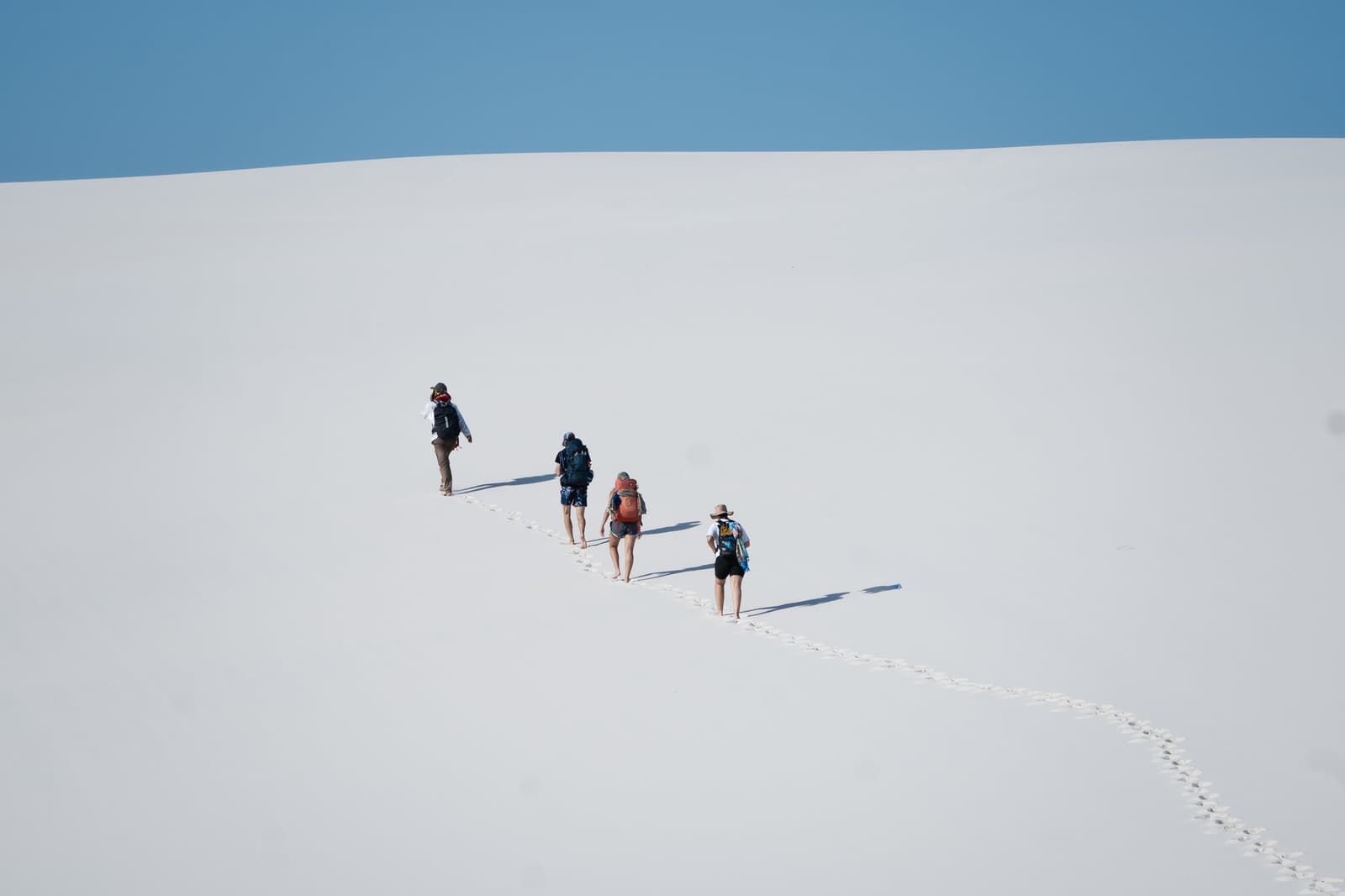 Vier rugzakken, één spoor, eindeloos wit zand voor je. Vier backpackers beklimmen achter elkaar een enorme witte zandduin tijdens een Lençóis Maranhenses-trip, met een spoor van blote voetafdrukken onder een heldere blauwe hemel