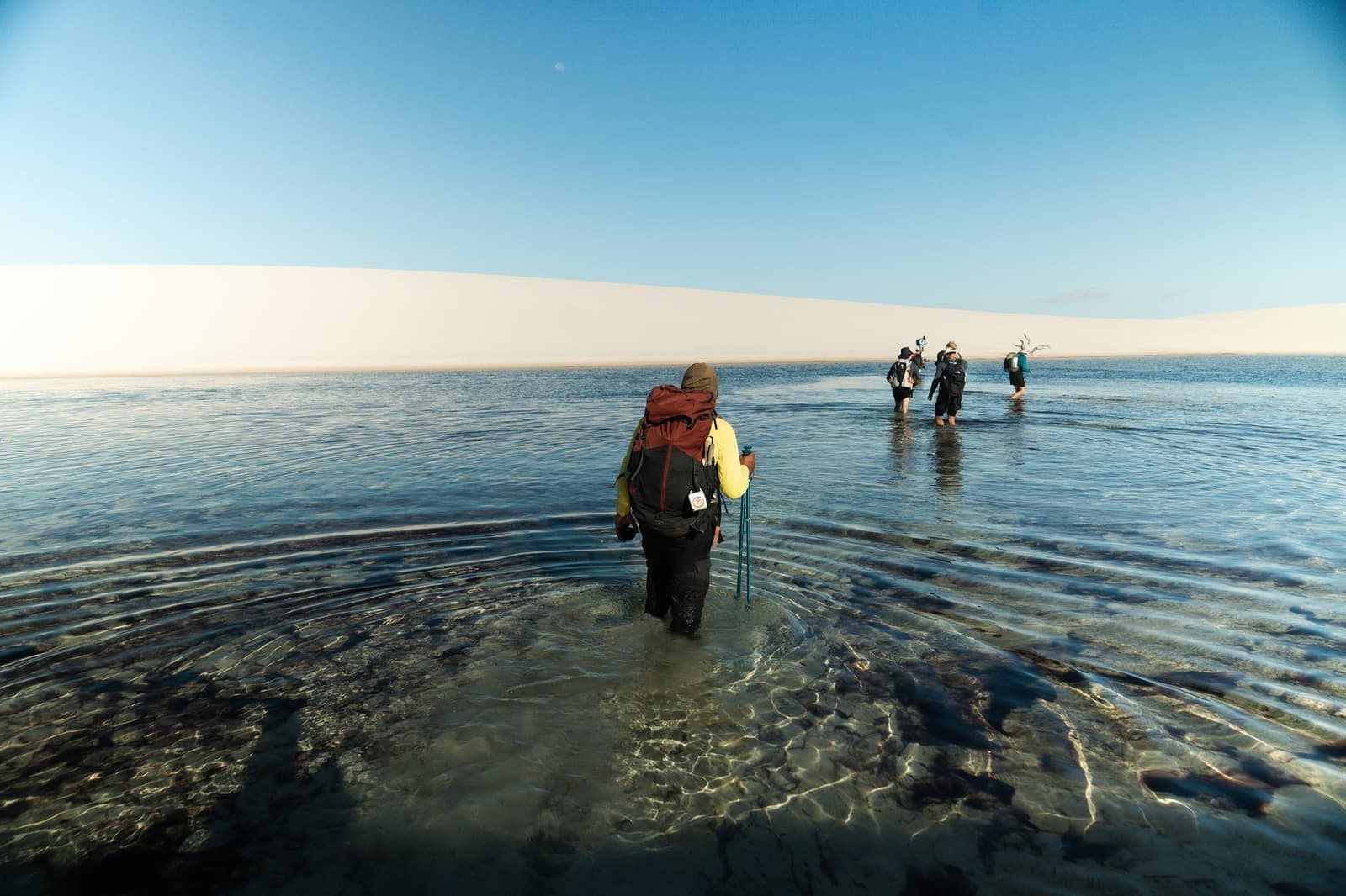 Sokken uit, rugzak op, de Lençóis speel je op zijn eigen voorwaarden. Een groep backpackers doorwaadt een heldere ondiepe lagune tijdens een Lençóis Maranhenses-reis, met uitgestrekte witte zandduinen aan de horizon onder een blauwe lucht