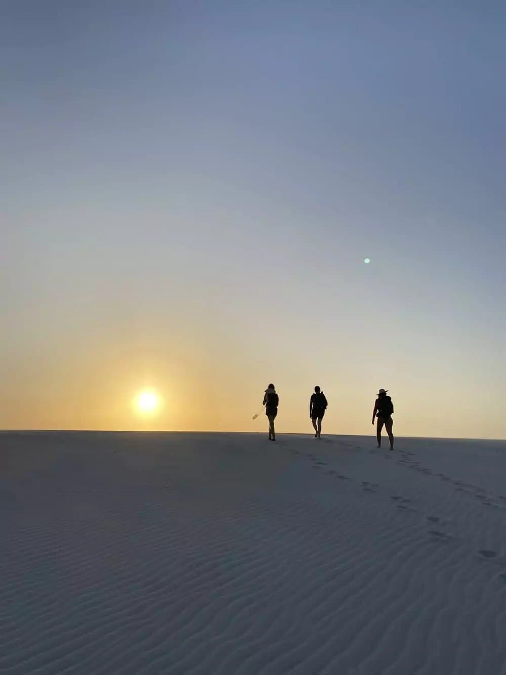 De duinen trekken zich niets aan van de tijd. De zon des te meer. Drie reizigers als silhouet tegen een gouden zonsonderganglucht op een uitgestrekte witte zandduin tijdens een Lençóis Maranhenses-trip, met voetafdrukken in het zand