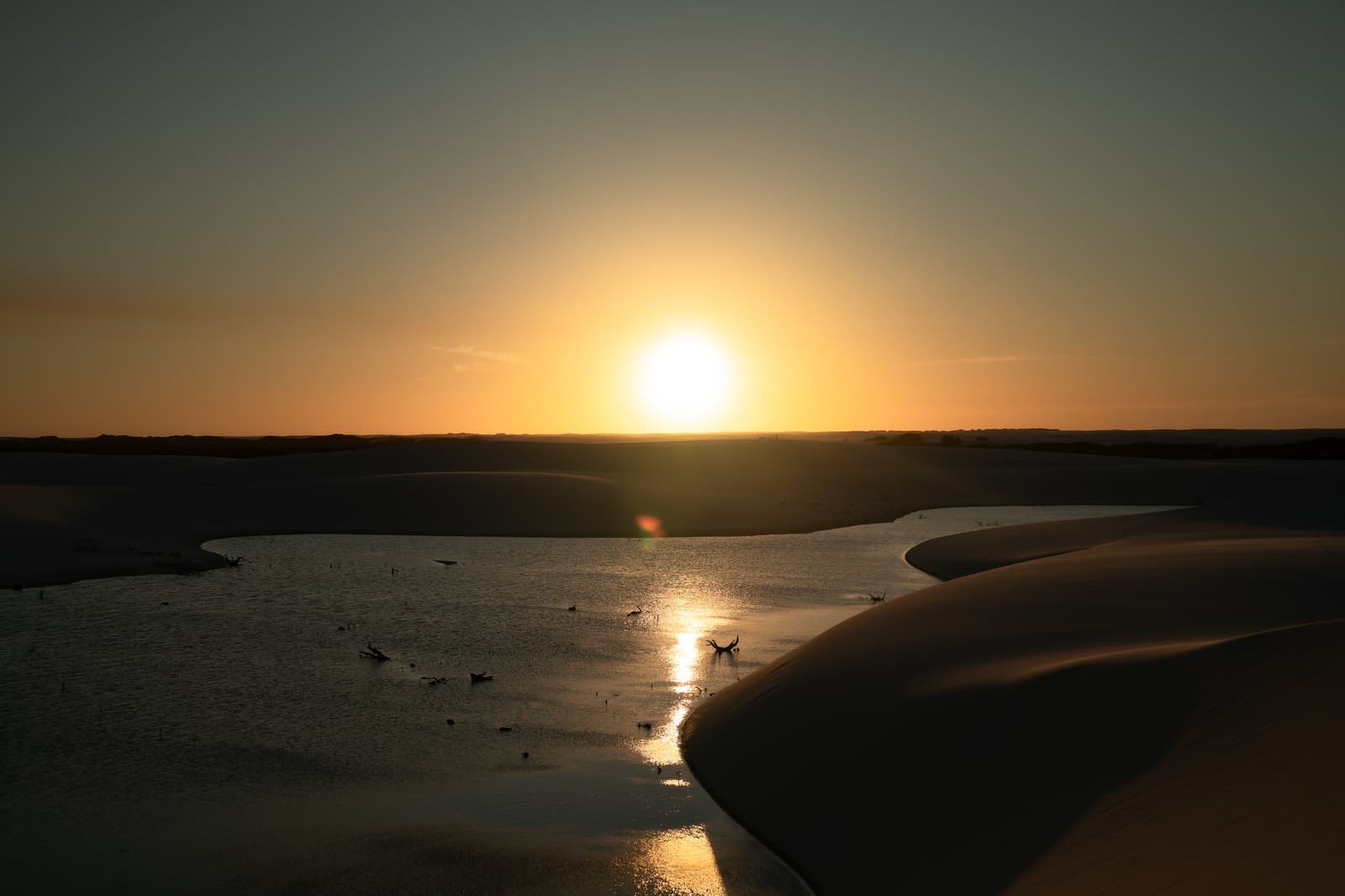 De duinen kleuren goud, de vogels pootjebaden, en verder is er weinig nodig. Ondergaande zon boven een ondiepe lagune tussen donkere zandduinen tijdens een Lençóis Maranhenses-trip, met vogels als silhouet op het spiegelgladde water