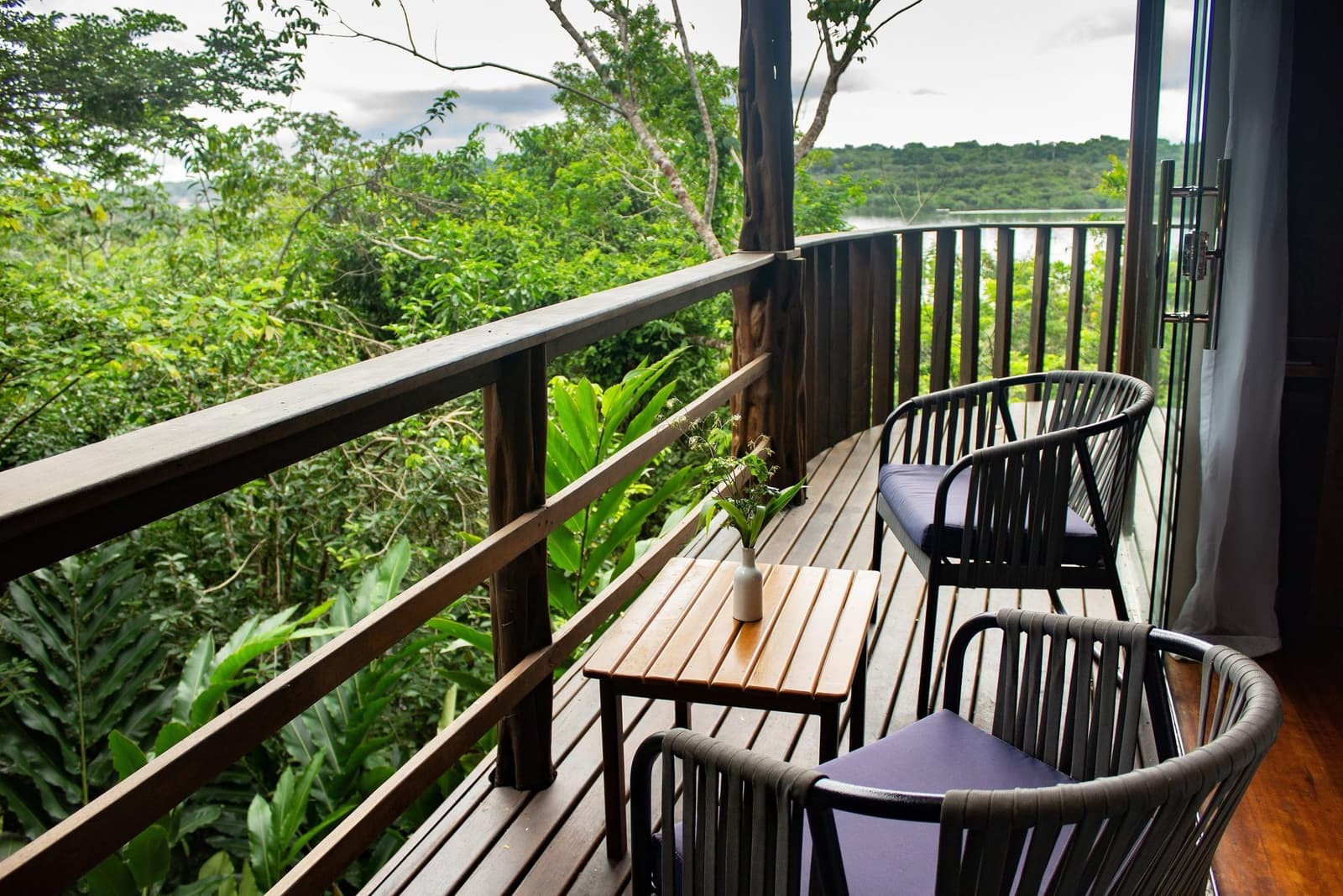 Hier schmeckt der Morgenkaffee einfach besser, der Regenwald macht das. Ein geschwungener Holzbalkon einer Luxuslodge im Amazonas mit zwei Rattanstühlen und einem kleinen Teakholztisch mit Blick auf das dichte grüne Regenwalddach und einen fernen Fluss