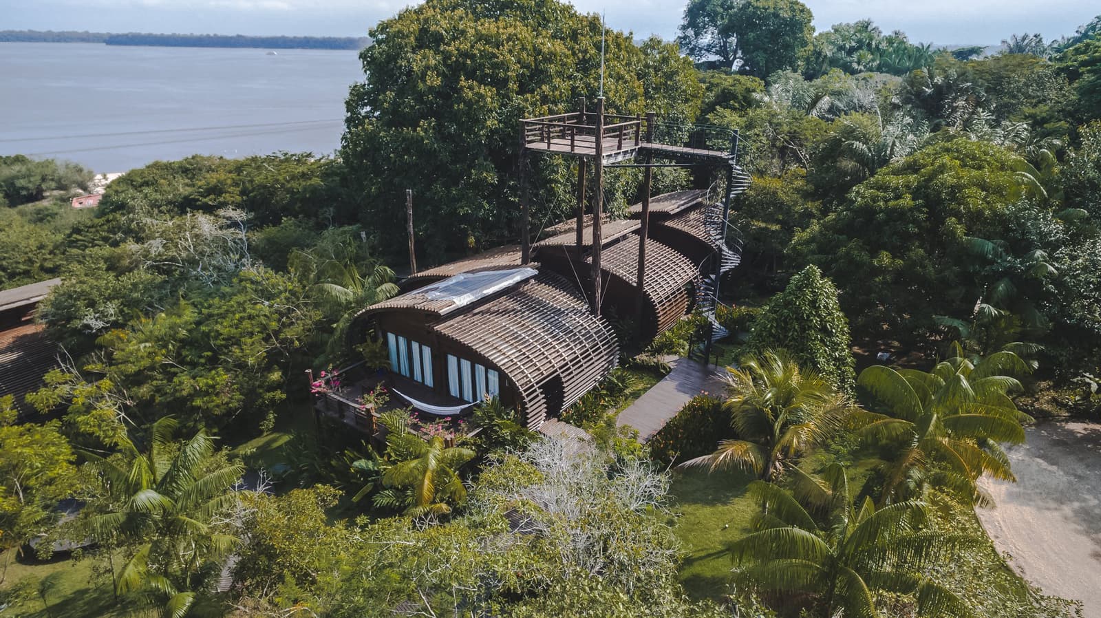 Ein Baumkronenweg auf dem Dach, die Lodge meint es ernst mit dem Vogelbeobachten. Luftaufnahme einer Luxus-Amazonas-Lodge in Brasilien mit einem geschwungenen Hartholzgebäude, einem lamellenverziertem Dach, einem Baumwipfelpfad und einem breiten Fluss im Hintergrund