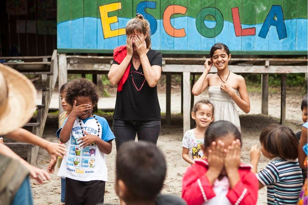 Die Kinder kennen die Regeln. Sie noch nicht. Genau das macht es so gut. Eine Reisende und Kinder aus der Gemeinde spielen ein Versteckspiel vor einem bunt bemalten Schulgebäude mit der Aufschrift 'ESCOLA' während eines Gemeindebesuchs in einem Luxus-Amazon-Lodge-Programm in Brasilien