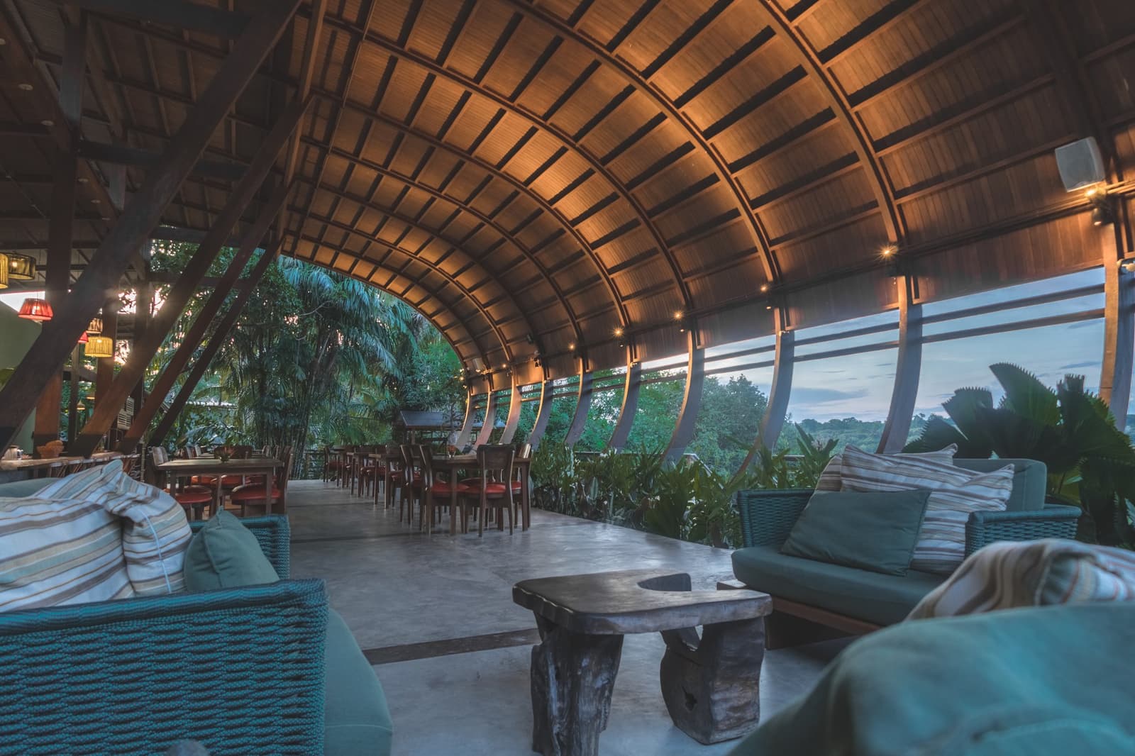 Hier setzt man sich kurz hin, und bleibt den ganzen Abend. Tonnengewölbte Holzdecke über der offenen Restaurantlounge einer Luxuslodge im Amazonas, mit Korbsofas, rustikalem Baumstamm-Couchtisch und raumhohen Panoramafenstern zum Regenwalddach in der Abenddämmerung