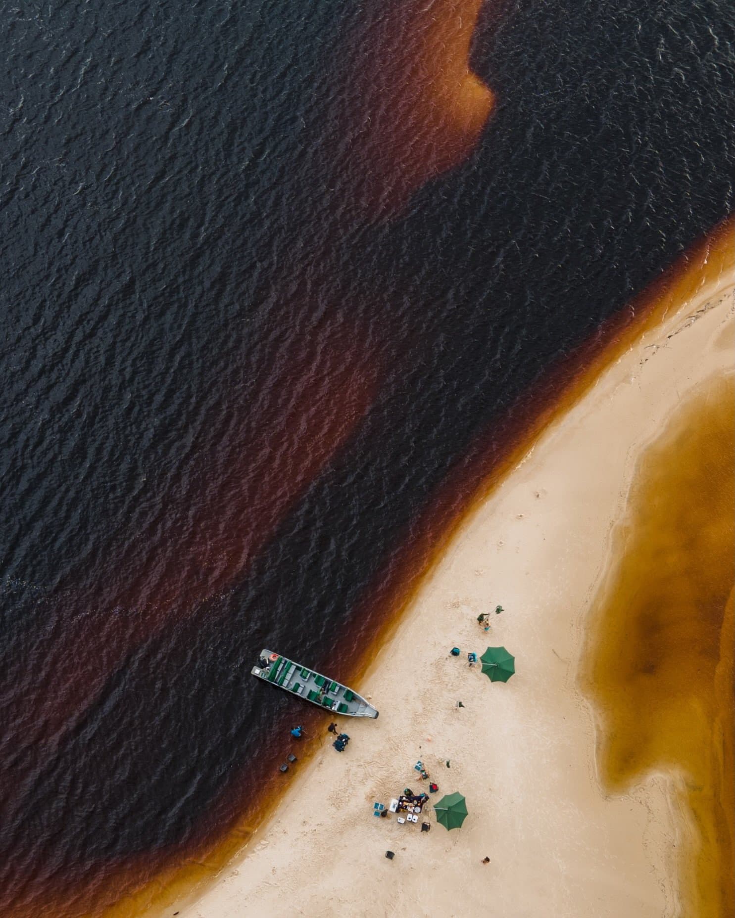 Zwei Flüsse, eine Sandbank, kein Grund zur Eile. Luftaufnahme eines kleinen Motorboots an einer weißen Sandbank neben dem dunkel-tanninhaltigen Fluss eines Luxus-Amazon-Lodges in Brasilien, mit zwei grünen Sonnenschirmen und einer Gruppe von Gästen