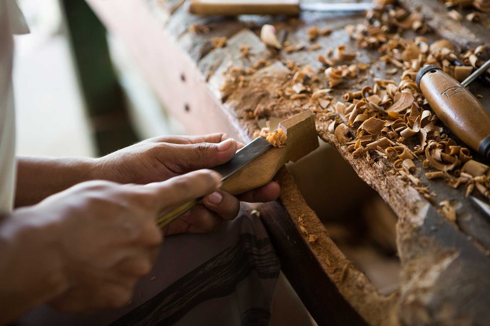 Geduldige Hände, scharfe Werkzeuge, eine Werkbank voller Geschichte. Nahaufnahme von Händen, die mit einem Stechbeitel ein Holzstück auf einer von frischen Holzspänen bedeckten Werkbank bearbeiten – lokales Kunsthandwerk in der Nähe einer Luxuslodge im brasilianischen Amazonas