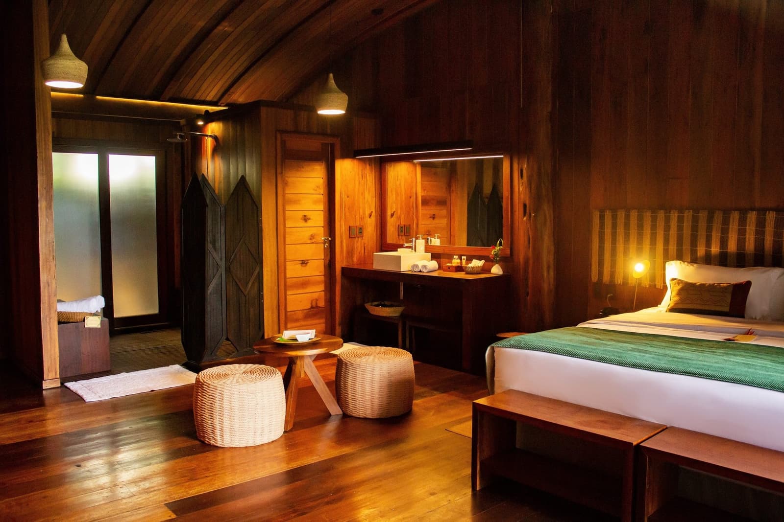 Holz, warmes Licht und der Regenwald direkt vor der Tür. Warmbeleuchtetes Holzkabinen-Interieur einer Luxus-Amazon-Lodge in Brasilien mit Doppelbett, Korbhockern, offener Waschtischanlage und glänzenden Hartholzböden unter einem gewölbten Holzdach