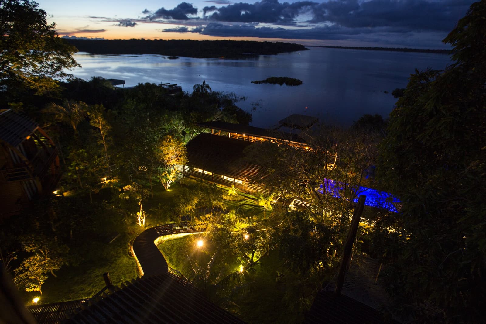 Die Lodge erwacht zum Leben, gerade wenn der Fluss sich in Orange taucht. Luftaufnahme in der Abenddämmerung einer Luxuslodge im Amazonas in Brasilien, mit warm beleuchteten Holzgebäuden, einem geschwungenen Steg und üppigem Dschungelgarten, der zu einem breiten dunklen Fluss mit bewaldeten Inseln unter dramatischem Abendhimmel führt