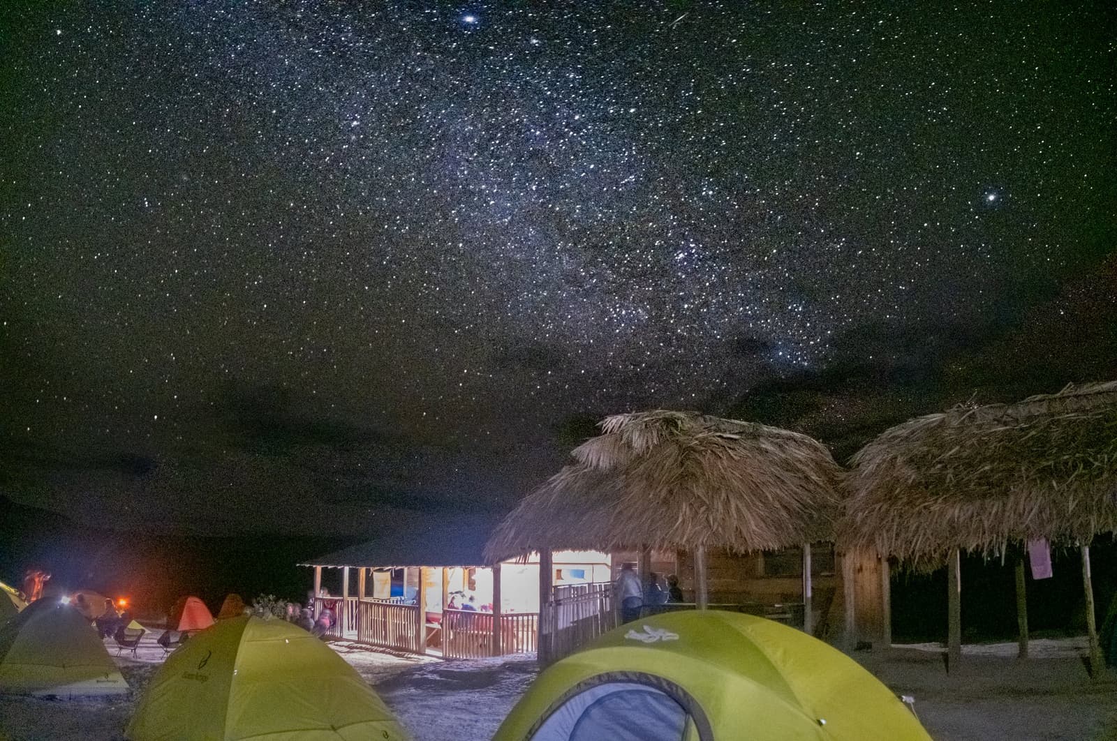 Your tent, a thatched hut, and half the galaxy overhead. Not bad for a Tuesday. Green dome tents and thatched-roof shelters at a remote campsite on the Mount Roraima trek, beneath a brilliant Milky Way filling the night sky