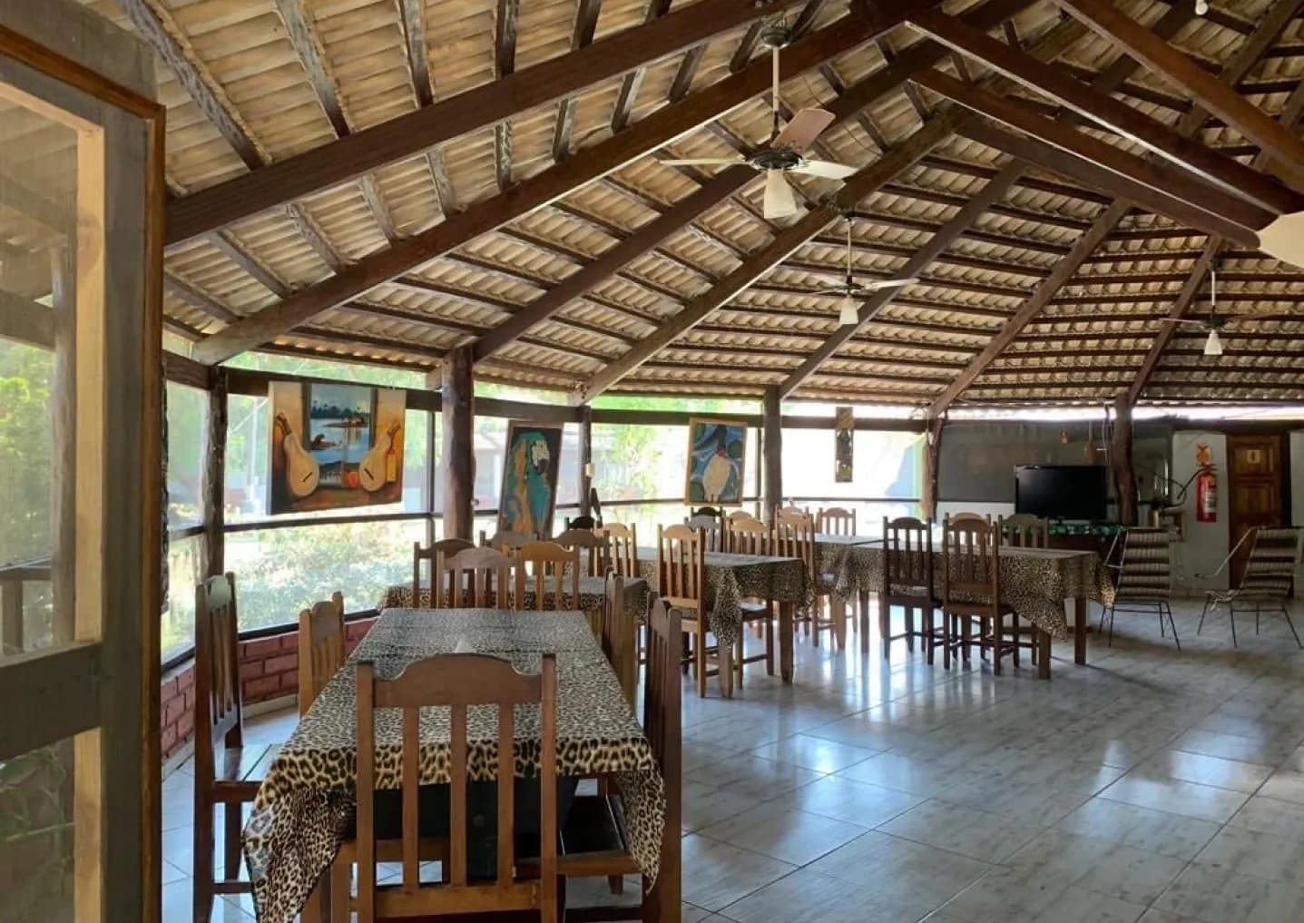 Rustikal, offen, mit einem Hauch Wildnis, genau das Richtige nach einer Tour. Speiseraum einer Lodge für Pantanal-Jaguar-Safaris mit Holzbalkendecke, Deckenventilator, Leopardenmuster-Tischdecken und Tiergemälden an der Fensterwand mit Blick ins Grüne