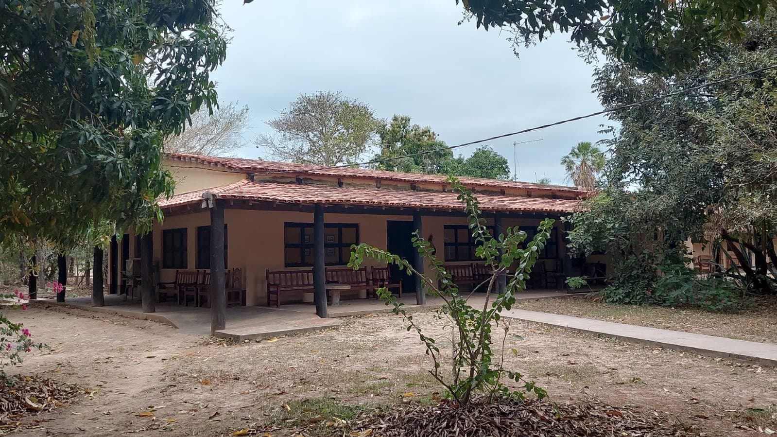 Kein Luxus, kein Schnickschnack, dafür Stille und guter Schlaf. Einstöckige Adobe-Lodge mit Terrakotta-Ziegeldach und überdachter Veranda mit Holzbänken, umgeben von alten Bäumen im Pantanal