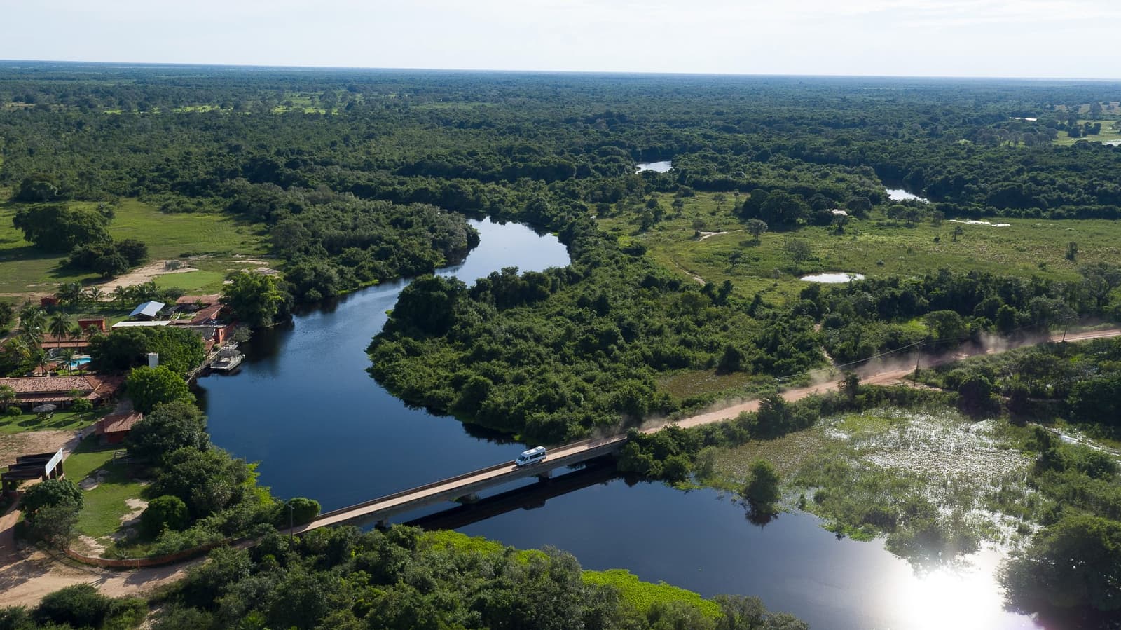 Eine kurze Überfahrt, und das Pantanal nimmt einen in Empfang. Luftaufnahme eines weißen Vans auf einer Betonbrücke über einen mäandernden Fluss, umgeben von dichter Pantanal-Feuchtgebietsvegetation und einer rustikalen Lodge am Ufer