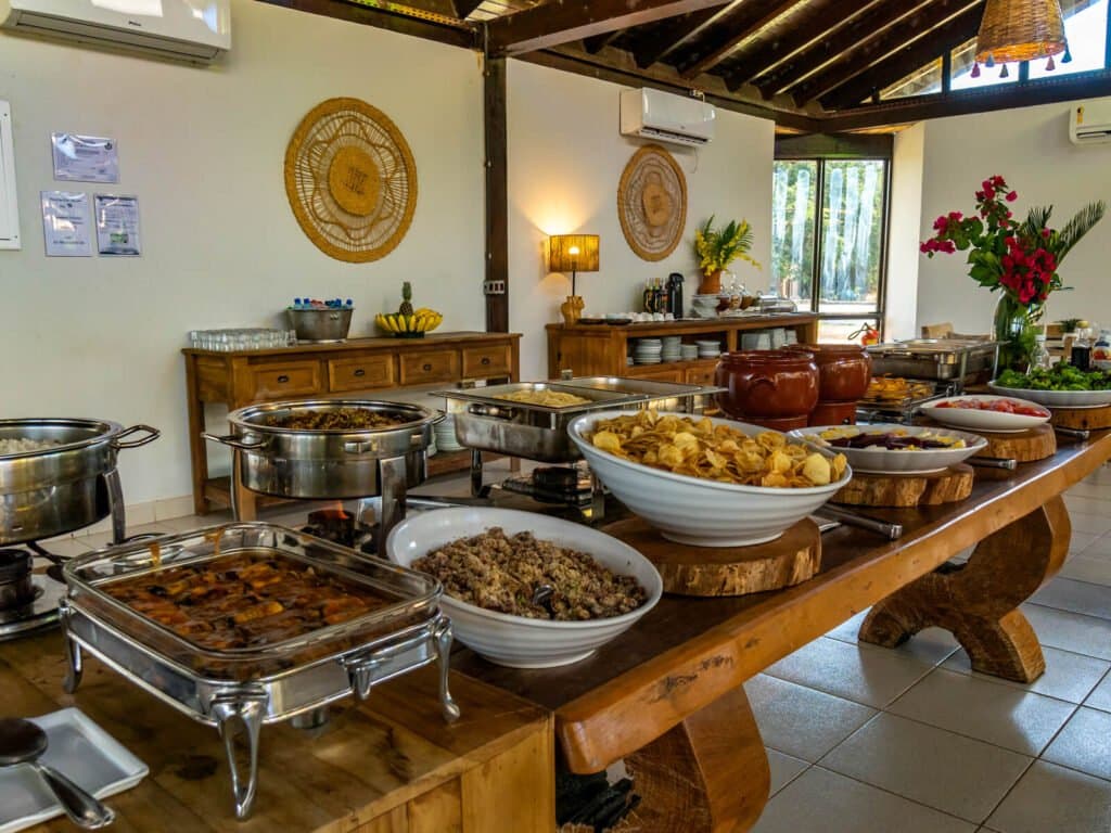 Regionale Küche, die nach einem Morgen auf dem Fluss stärkt. Ein reichhaltiges brasilianisches Buffet auf einem rustikalen Holztisch in einer Lodge beim Pantanal-Jaguar-Safari in Porto Jofre, mit Warmhaltebehältern, Farofa, Snacks und frischen Blumen