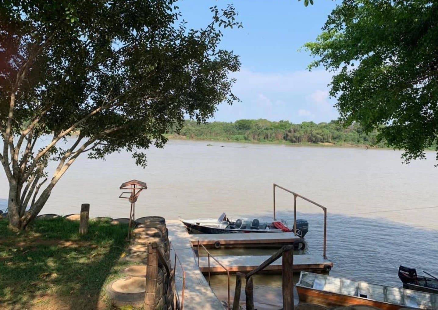 Boote bereit, der Fluss wartet, der Aufbruch ins Pantanal beginnt hier. Ein hölzerner Flusssteg mit vertäuten Motorbooten an einem breiten, braunfarbenen Fluss, umrahmt von Uferbäumen an einem sonnigen Tag