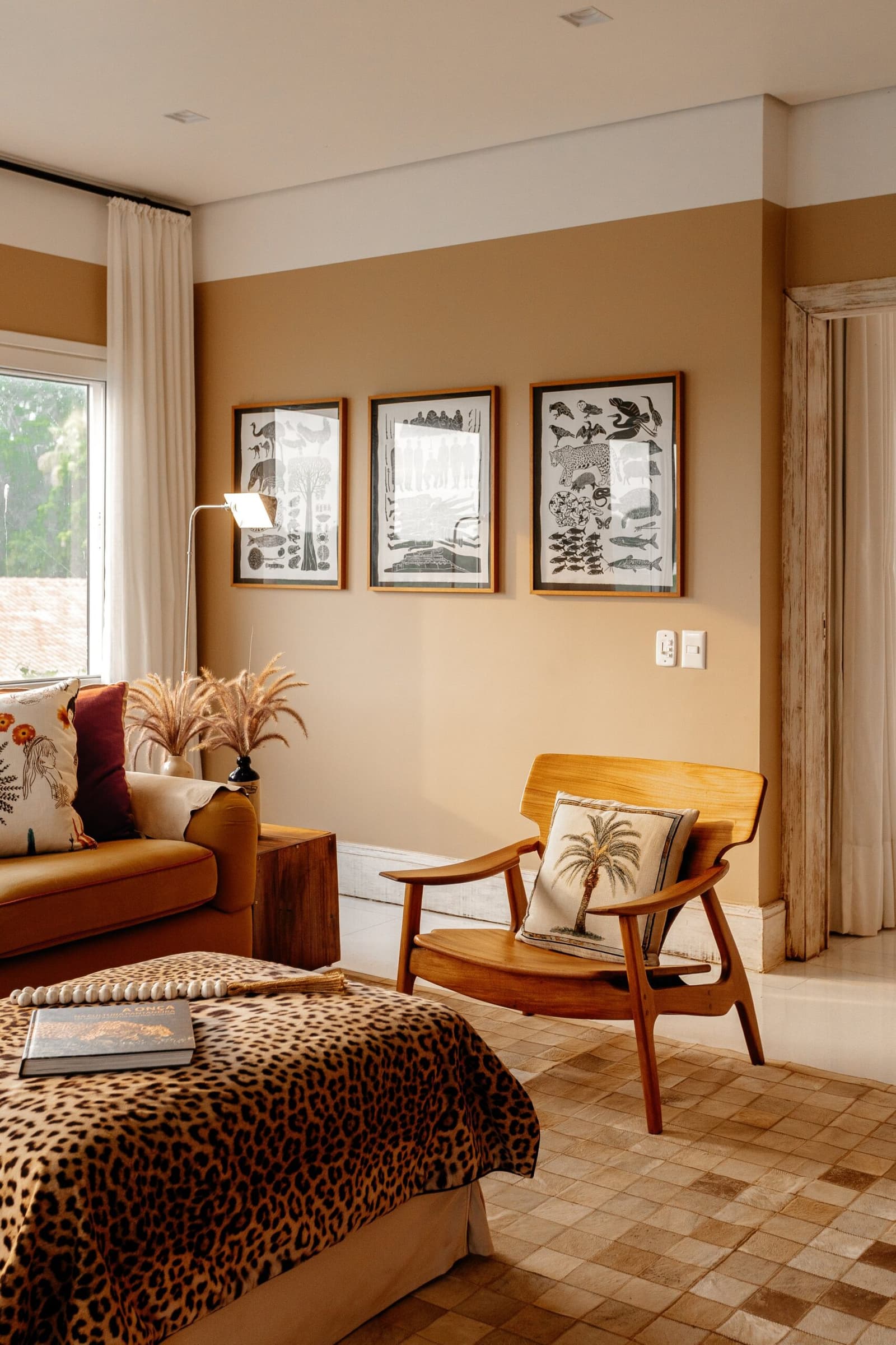 The kind of room that makes you forget you packed field clothes. A warmly decorated suite at Caiman Ecological Refuge featuring a leopard-print bedspread, mid-century timber armchair with a palm-motif cushion, and three framed wildlife illustrations on ochre walls — embodying pantanal luxury safari style