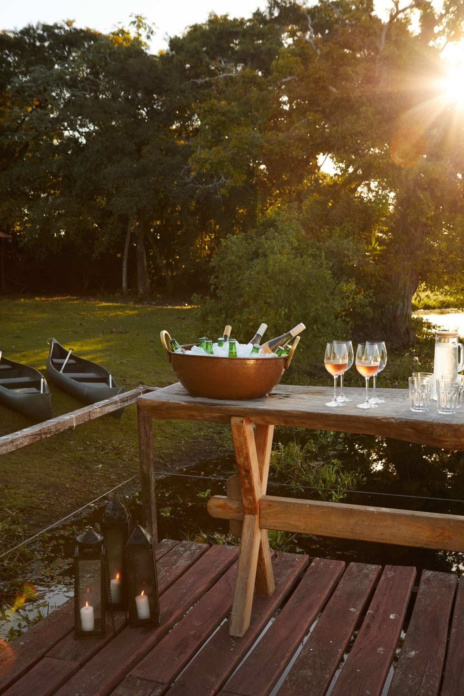Canoes done for the day, drinks very much not. A copper ice bucket filled with chilled bottles and rosé wine glasses on a rustic wooden table at sunset on a riverside deck, with canoes on the bank and candlelit lanterns below — part of the pantanal luxury safari experience at Caiman