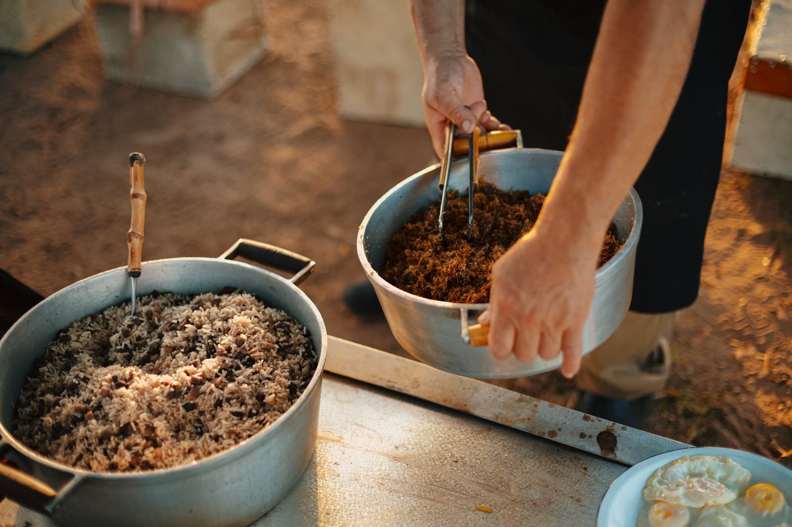 Camp food done right: honest, hearty, and ready before the sun sets. Hands stirring seasoned shredded meat in an aluminium pot beside a large pot of rice and black beans, served outdoors as part of a pantanal luxury safari all-inclusive meal