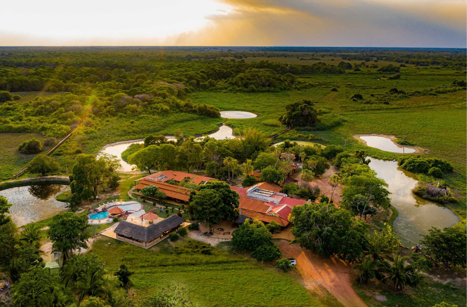 Je slaapplaats ligt midden in het wetland, niet ernaast. Luchtfoto van een Pantanal eco-lodge omgeven door kronkelende lagunes, weelderige moerasvegetatie en een hoofdgebouw met terracottadaken en een zwembad bij gouden zonsondergang