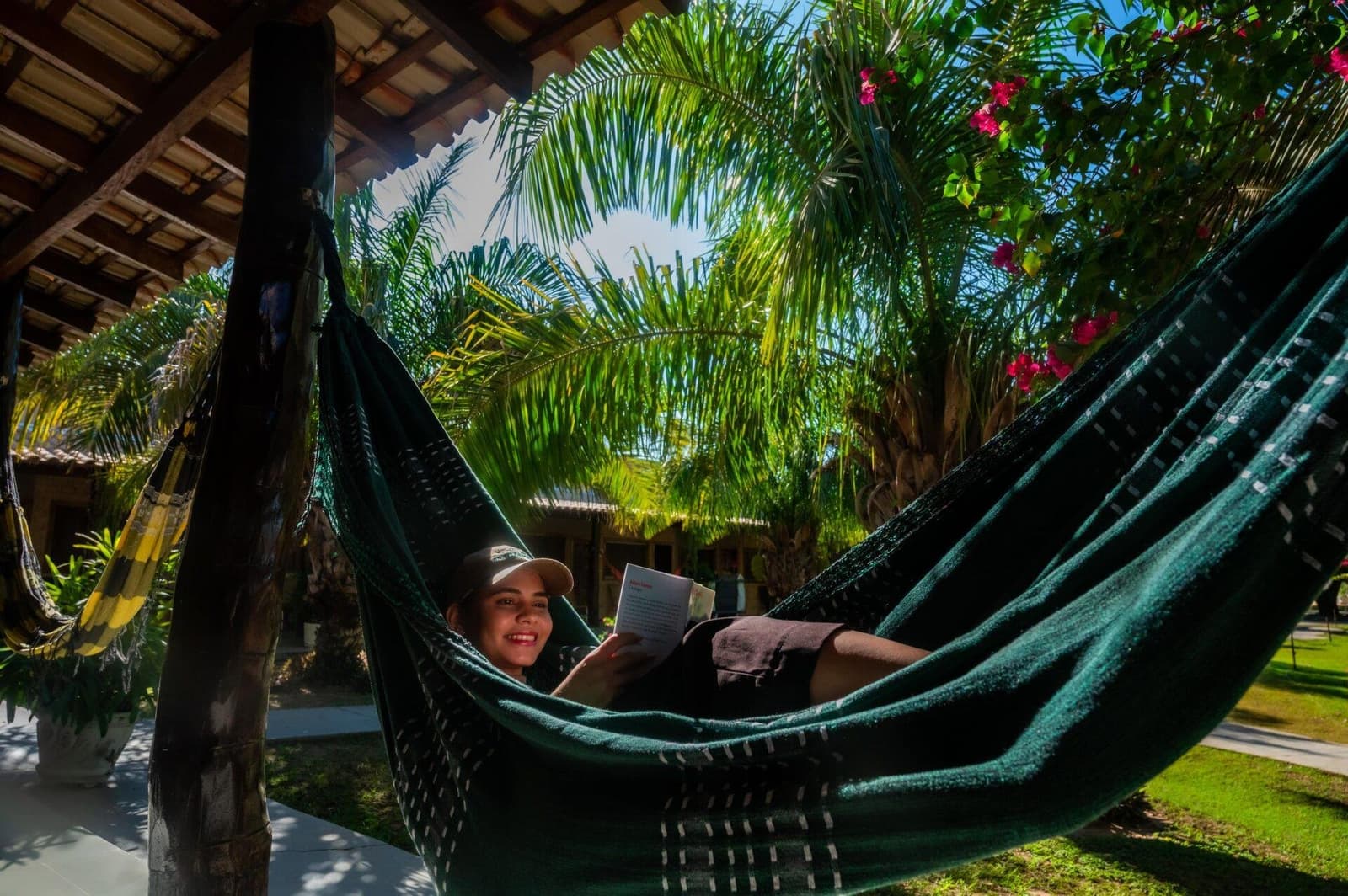 Tussen twee safariritten door verdient een goed boek zijn plek. Een glimlachende reizigster leest een boek in een donkergroene hangmat op de beschaduwde veranda van een lodge, omgeven door palmtakken en roze bougainvillea, tijdens een Pantanal-tour vanuit Cuiabá