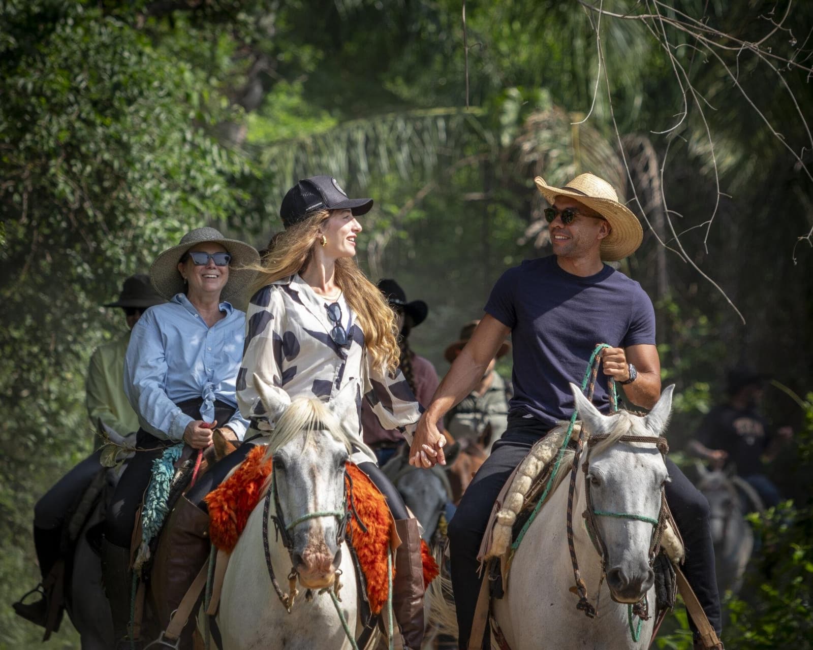 Hand in hand op de rug van een paard, ergens in de Pantanal. Werkt altijd. Een groep lachende ruiters op grijze paarden rijdt door een weelderig groen pad tijdens een Pantanal-tour vanuit Cuiabá, met vooraan een stel dat elkaars hand vasthoudt in het zadel