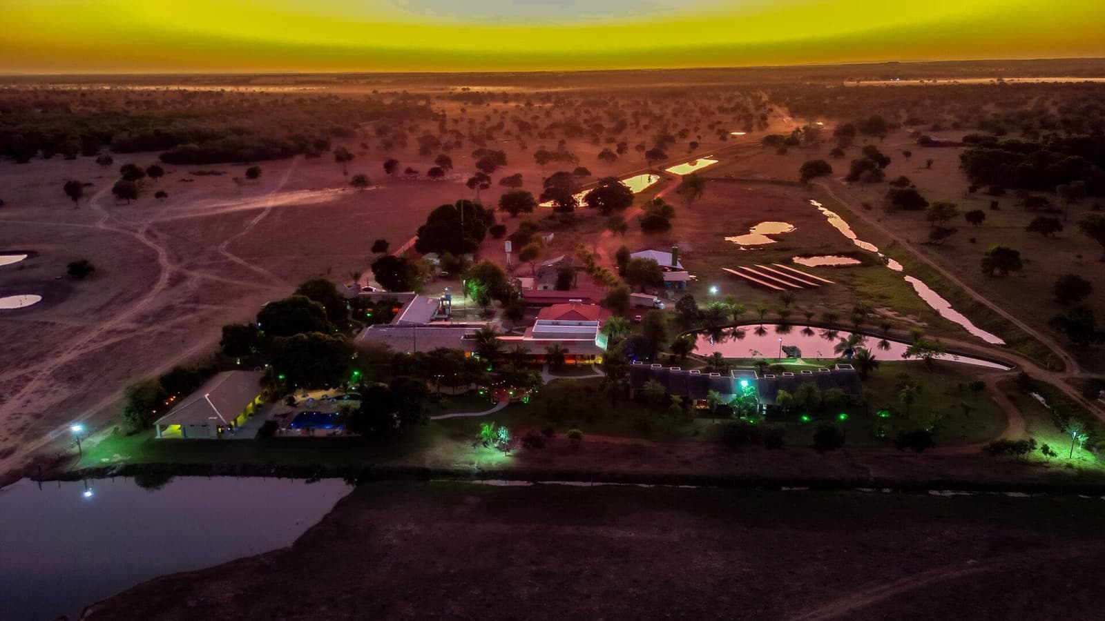 Als de zon vertrekt, neemt de lodge het over met eigen licht. Luchtvogelperspectief van een verlichte Pantanal-lodge bij zonsondergang, omringd door moeraspoelen en open struikland tot aan de horizon onder een levendige geel-oranje lucht