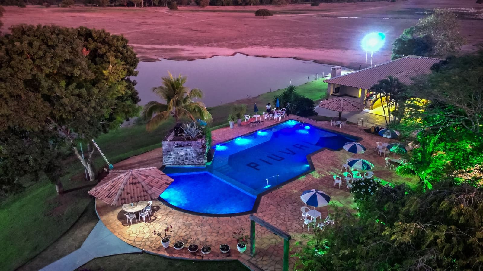 Een dag vol wilde dieren eindigt het best met dit uitzicht en een koele duik. Luchtfoto bij nacht van een verlicht zwembad bij een lodge voor de Pantanal-tour vanuit Cuiabá, met terrasparasols, een stenen waterornament en een natuurlijk meer voor een open savannelandschap