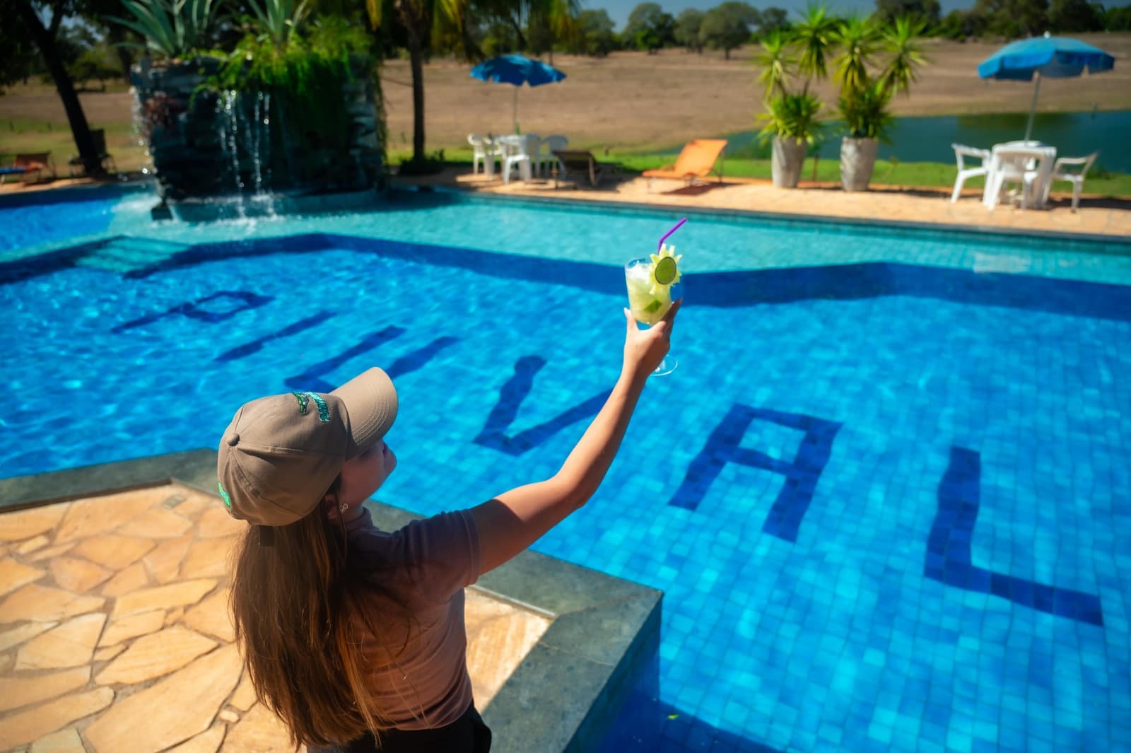 Welkom in het Pantanal. Cocktail eerst, wildlife daarna. Een gast heft een limoencocktail bij het blauw betegelde zwembad van een Pantanal-lodge vanuit Cuiabá, met ligbedden en een rustig meer op de achtergrond