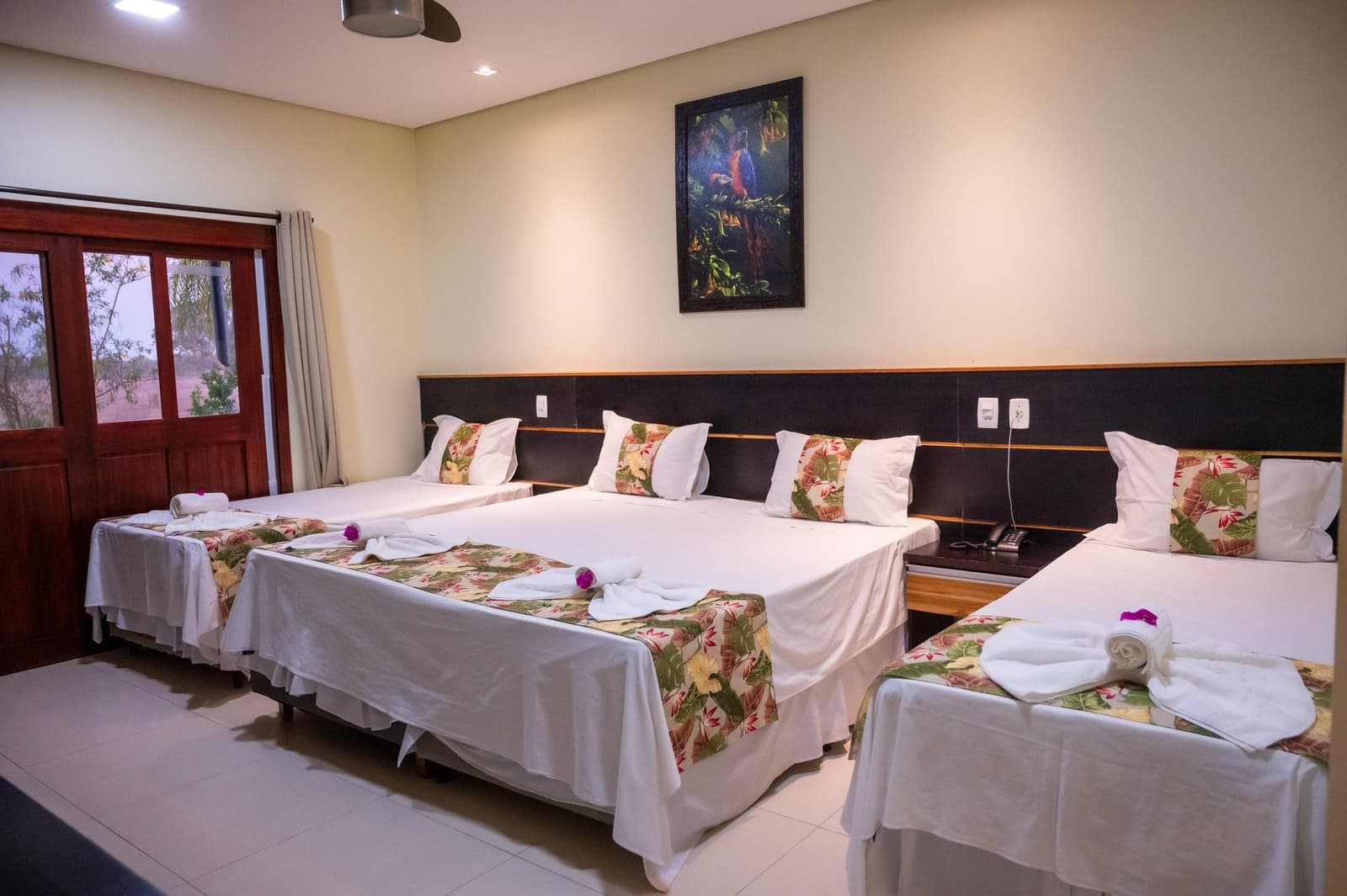 Na een dag vol wilde dieren smaakt een opgemaakt bed extra goed. Driepersoonshotelkamer in een lodge voor een Pantanal-tour vanuit Cuiabá, met drie opgemaakte bedden met tropische bloemenloper, opgerolde handdoeken met paarse orchideeënaccenten en een ingelijst schilderij van een ara boven het hoofdbord