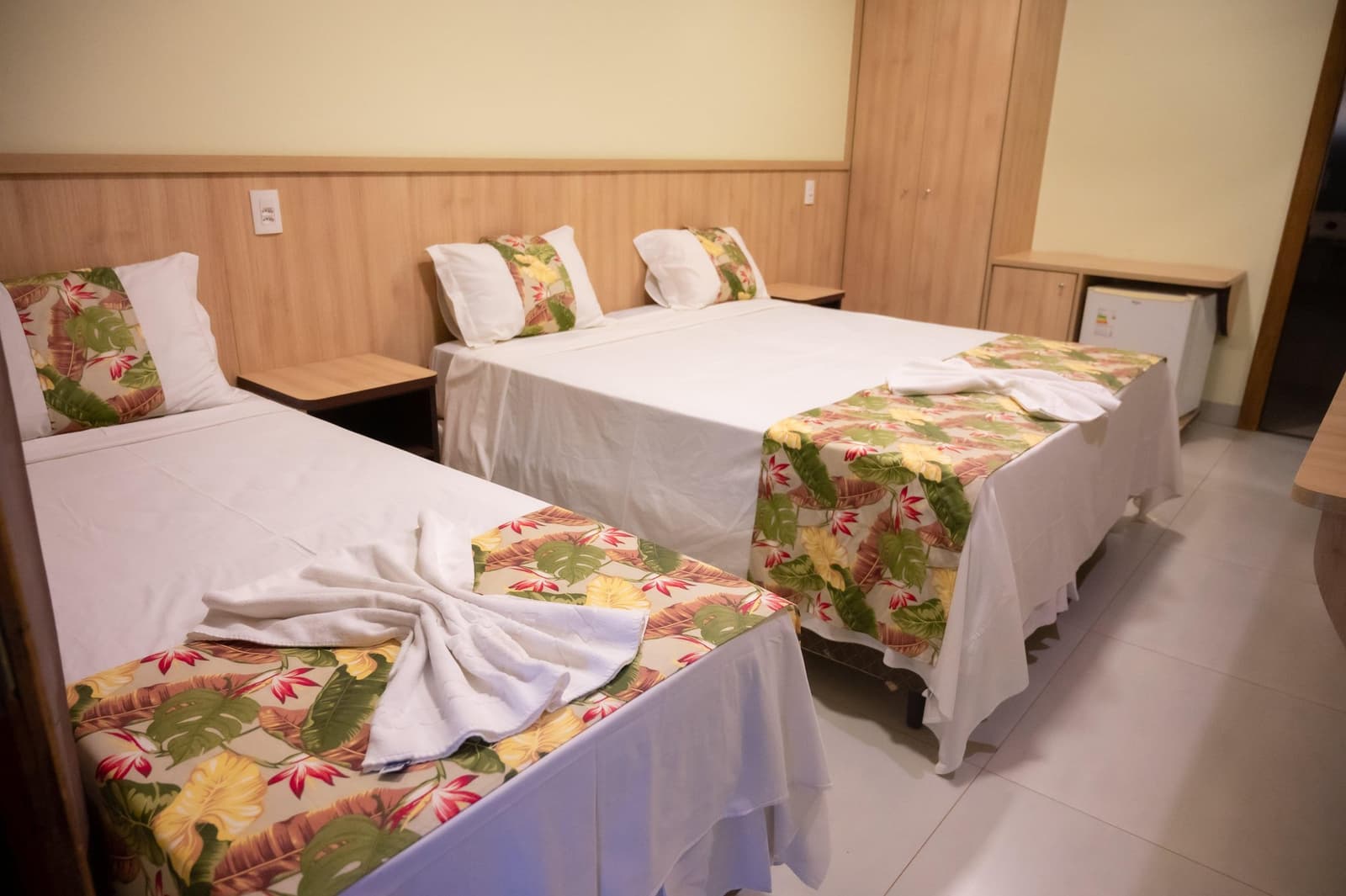 Strakke bedden, een gevouwen handdoek en een tropische print, iemand hier neemt het welkom serieus. Drie opgemaakt bedden met tropische bloemensprei en gevouwen witte handdoeken in een houten gastenkamer tijdens een Pantanal Tour vanuit Cuiabá