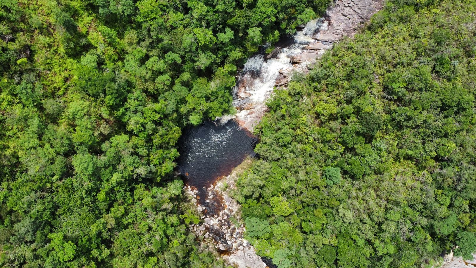 A trilha não avisa, mas a paisagem compensa na hora. Vista aérea de uma cachoeira em cascata sobre rocha exposta despejando água em uma piscina natural escura com taninos, cercada por densa cobertura florestal na trilha do Vale do Pati