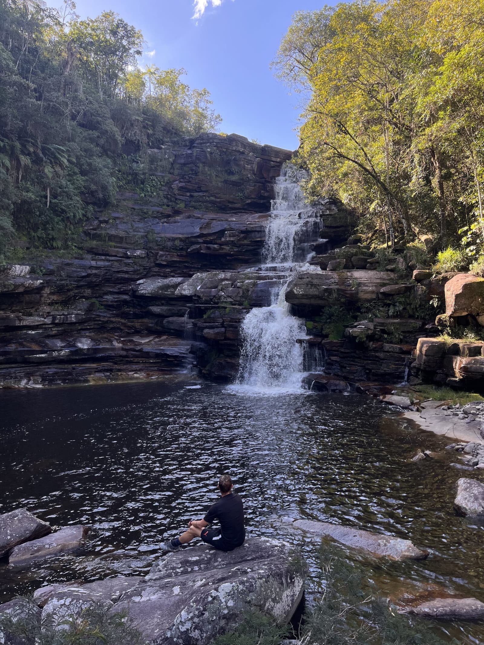 Tem lugares que pedem silêncio, e você entende na hora. Um caminhante sentado sozinho sobre uma rocha plana de arenito à beira de um poço natural escuro contempla uma cachoeira em degraus caindo por paredões rochosos estratificados durante a trilha do Vale do Pati, cercado por mata verde sob céu azul