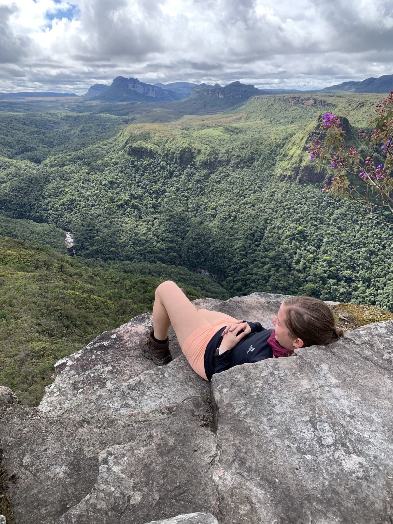 O melhor mirante da trilha não precisa de placa para ser encontrado. Uma caminhante deitada sobre uma borda rochosa contempla o profundo canyon verde do Vale do Pati com montanhas de arenito de topo plano e uma cachoeira ao fundo sob céu nublado