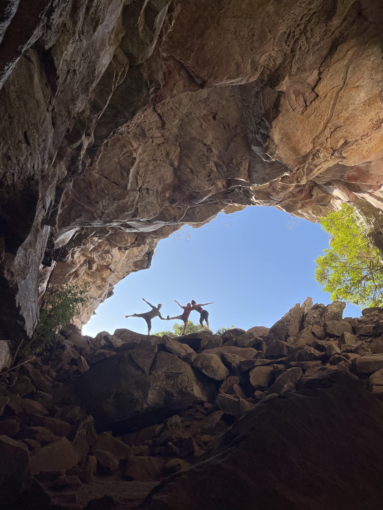 A gruta deu o cenário; a coreografia foi por conta deles. Dois caminhantes em poses de equilíbrio sobre pedras emolduradas por um imenso arco de arenito em uma gruta durante a trilha do Vale do Pati, céu azul e copas de árvores visíveis pela abertura