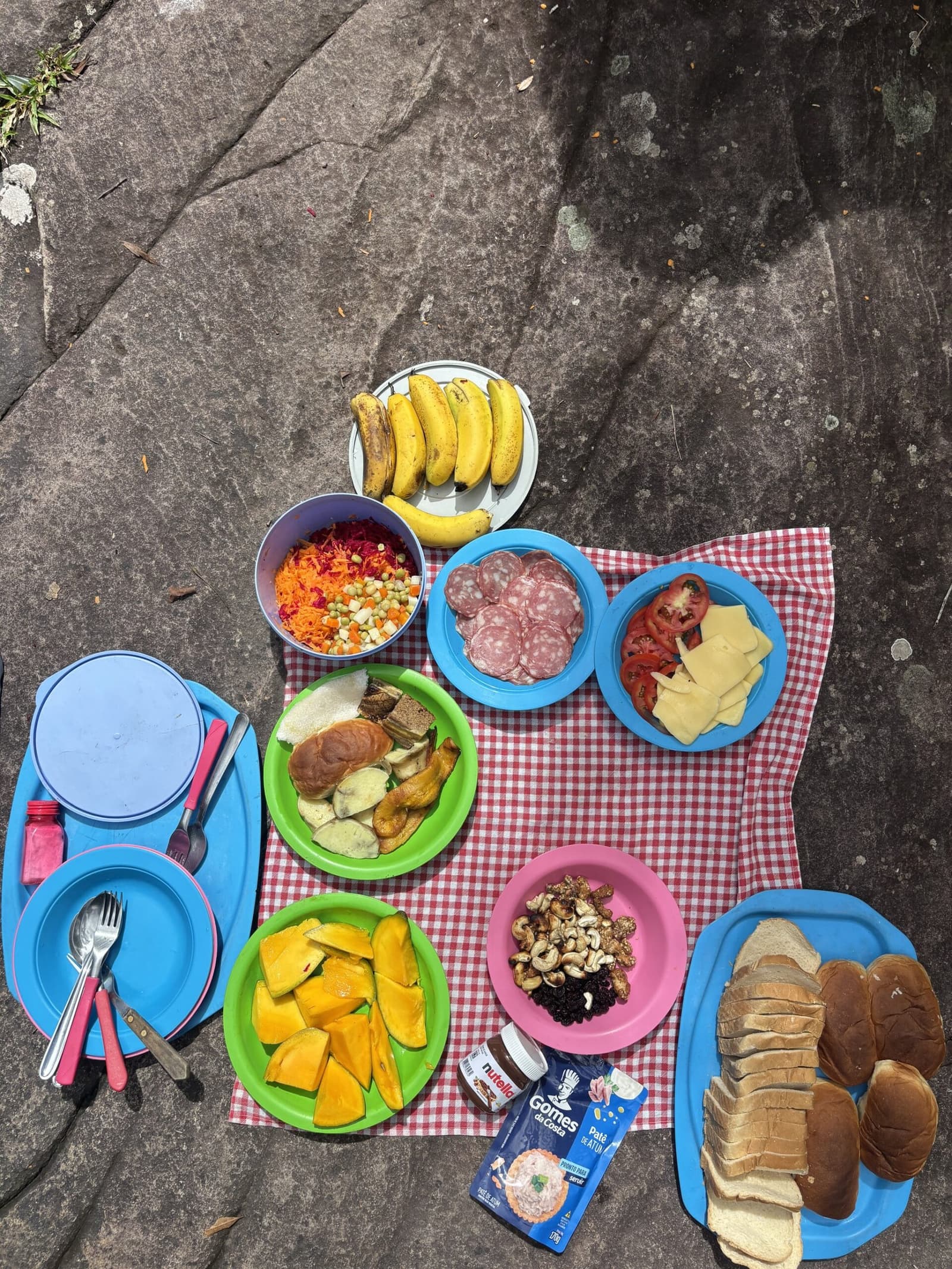 Mesa posta na pedra, e ninguém reclamou do cardápio. Pratos coloridos com frutas, salaminho, queijo, legumes ralados, castanhas e pão fatiado dispostos sobre uma toalha xadrez vermelha e branca em cima de uma pedra durante o almoço na trilha do Vale do Pati
