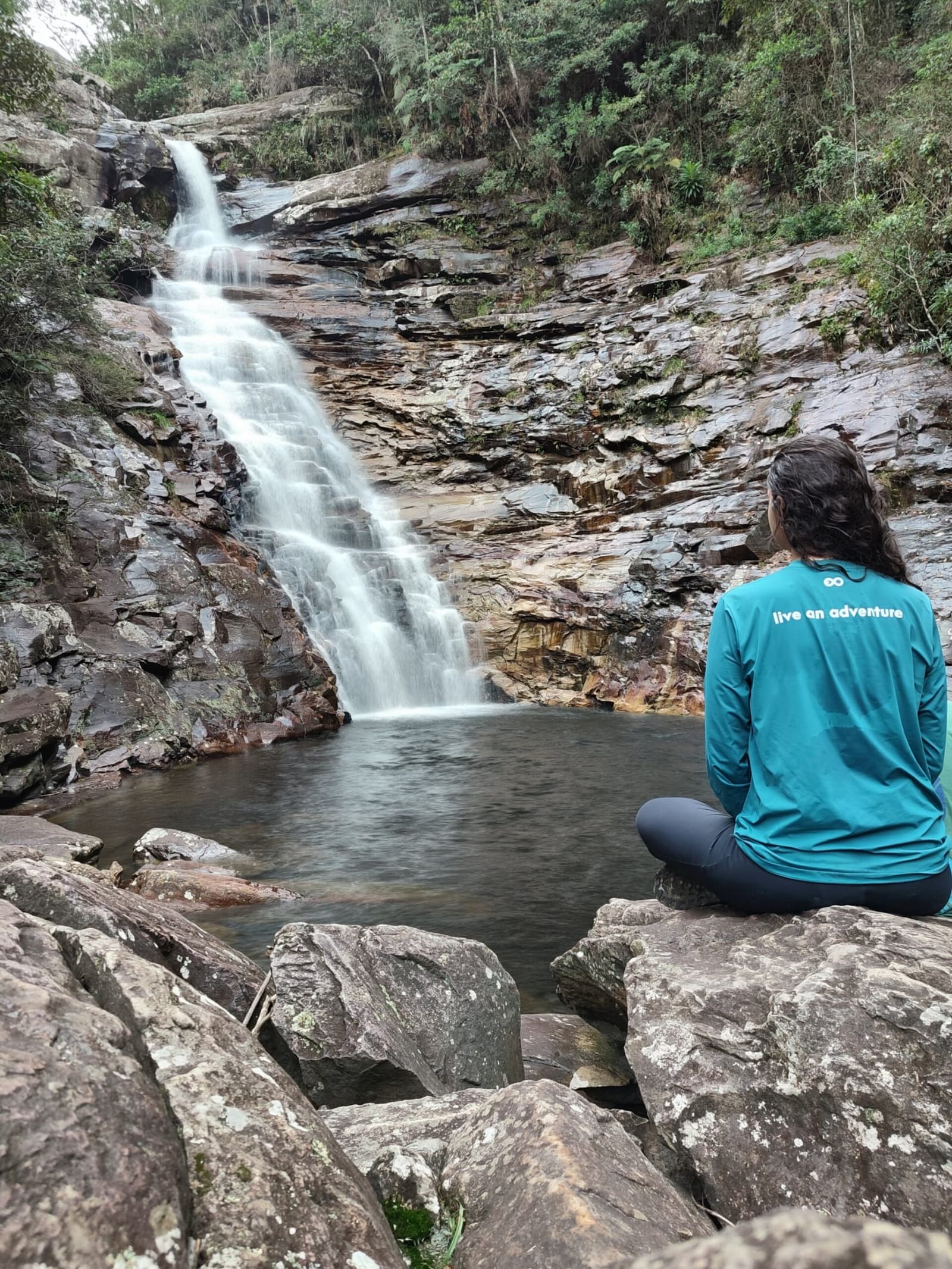 A cachoeira não vai a lugar nenhum. Você também não precisa. Uma caminhante de blusa verde-azulada senta de pernas cruzadas sobre pedras e contempla uma cachoeira que desce suavemente por paredões rochosos em camadas até uma piscina natural cercada de floresta